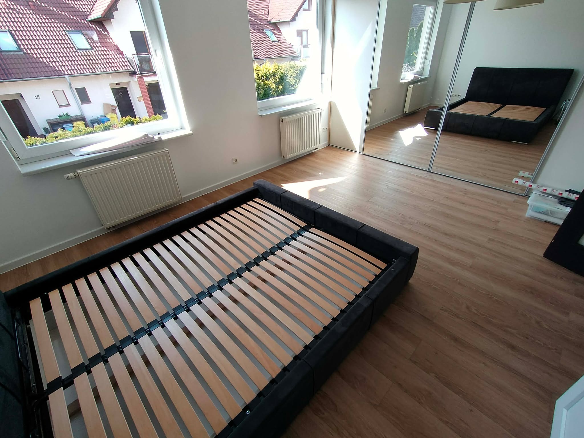 Jasne wnętrze z drewnianą podłogą i częściowo złożonym łóżkiem. Widok na okno z budynkami na zewnątrz. W tle lustrzana szafa odbijająca fragment pokoju.