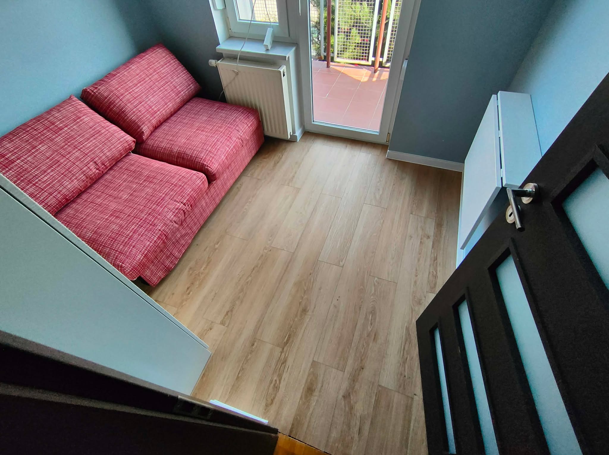 Jasne panele podłogowe w pokoju z wyjściem na balkon, widoczne z perspektywy otwartych drzwi. Czerwona sofa i grzejnik przy ścianie. Ujęcie z góry.