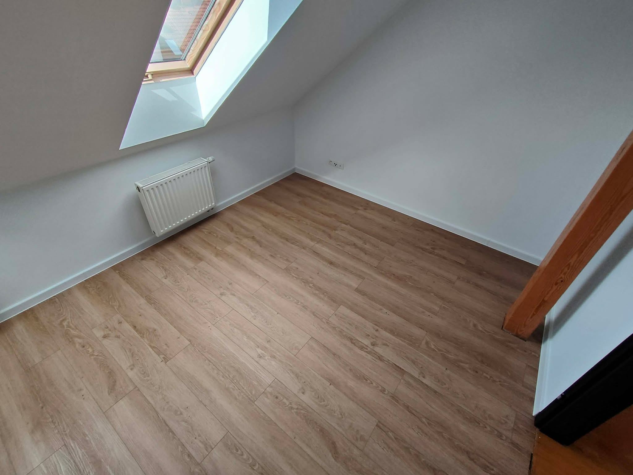 Jasne panele podłogowe w pokoju na poddaszu z oknem dachowym i grzejnikiem. Widoczny fragment drewnianej belki konstrukcyjnej.