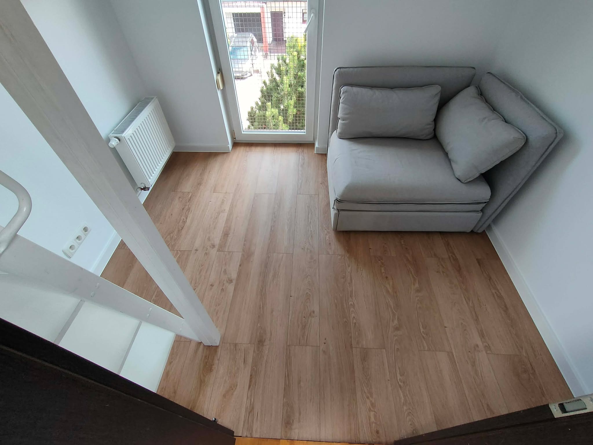 Jasne panele podłogowe w pokoju z białymi ścianami, widok z góry. Fragment schodów, okno z widokiem na zieleń i szara sofa w rogu.