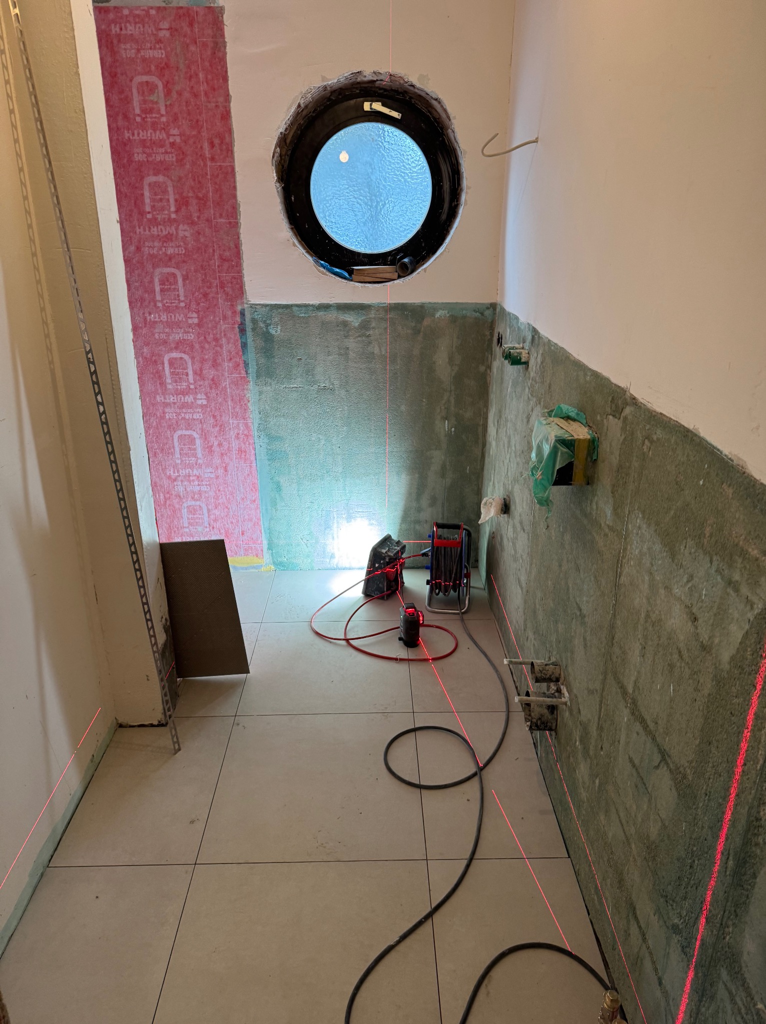 Łazienka w trakcie prac wykończeniowych: położone płytki podłogowe, ściany przygotowane pod glazurę, widoczny laser krzyżowy i materiały budowlane. Okrągłe okno.