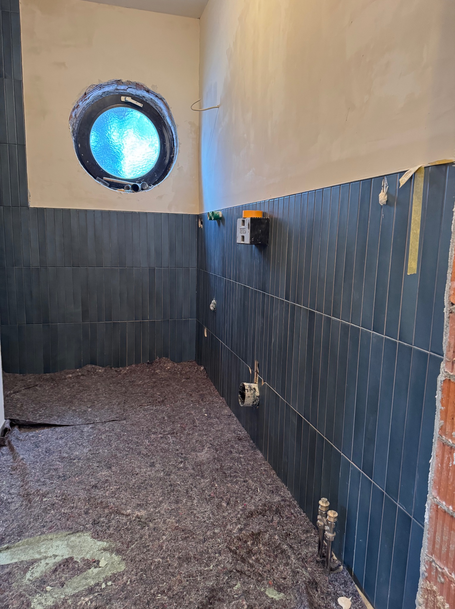 Niebieskie, pionowe płytki w łazience z okrągłym oknem. Podłoga zabezpieczona. Widoczne instalacje i fragment ściany z cegły. Etap prac wykończeniowych.