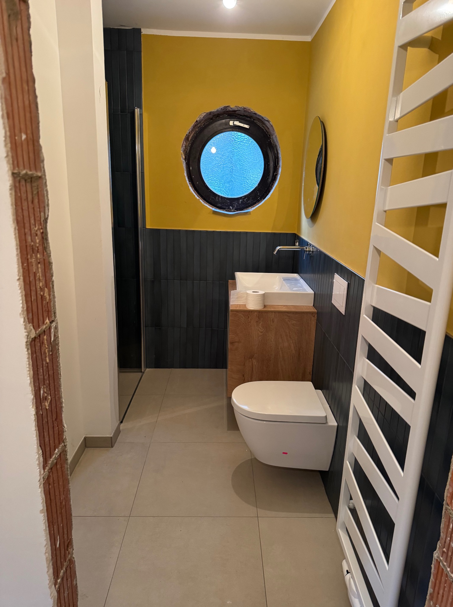 Żółto-czarna łazienka z okrągłym oknem i grzejnikiem drabinkowym. Fragment ściany w stanie surowym. Umywalka z szafką i podwieszana toaleta.