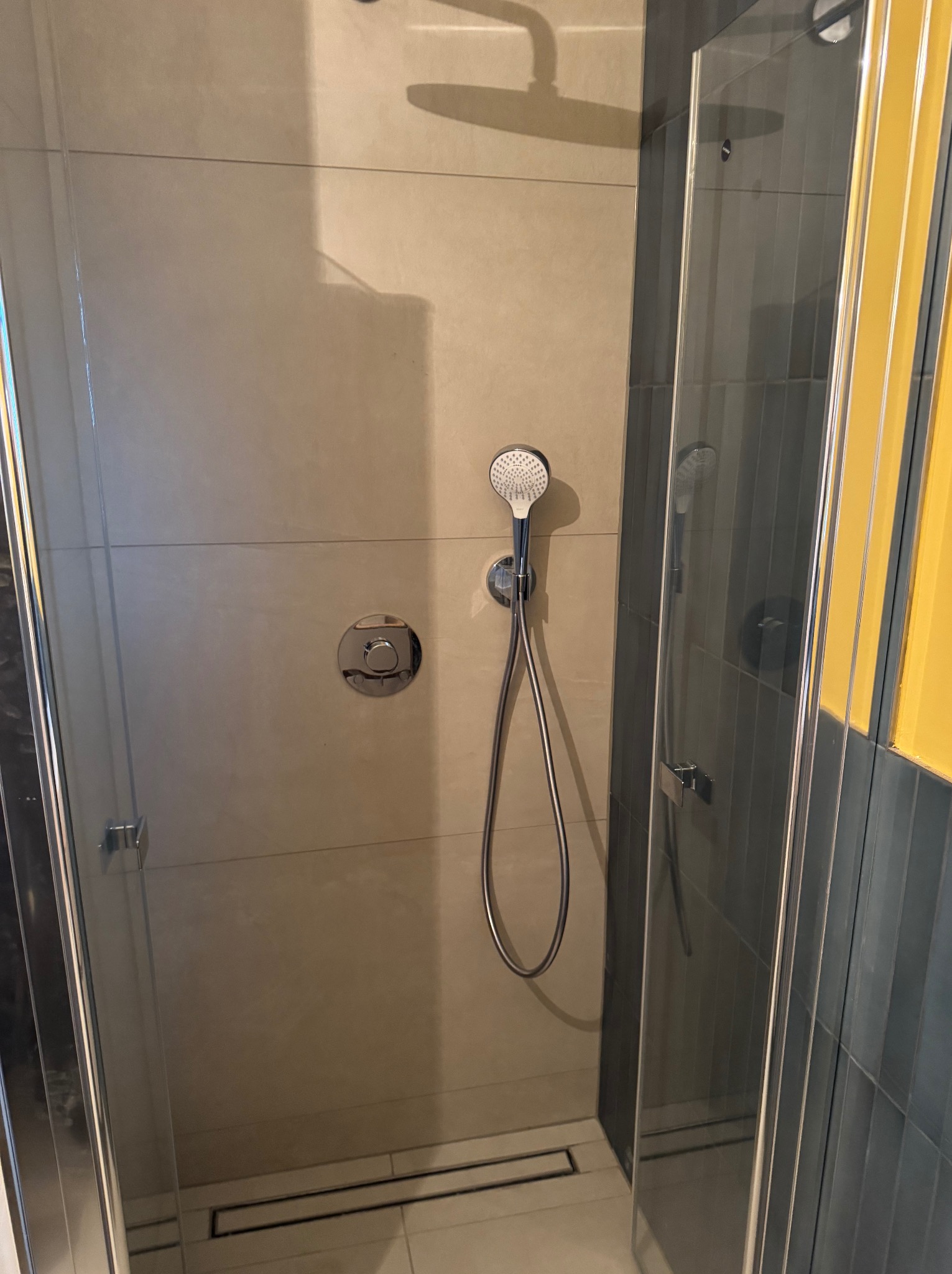 Nowoczesna kabina prysznicowa z deszczownicą i szklanymi drzwiami. Beżowe płytki na ścianach, chromowana armatura. Widoczny odpływ liniowy w podłodze.