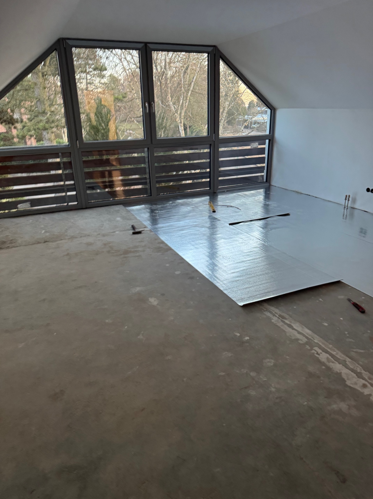 Poddasze z oknami i częściowo ułożoną folią izolacyjną na podłodze. Widoczne narzędzia i surowy beton. Przygotowanie do dalszych prac wykończeniowych.