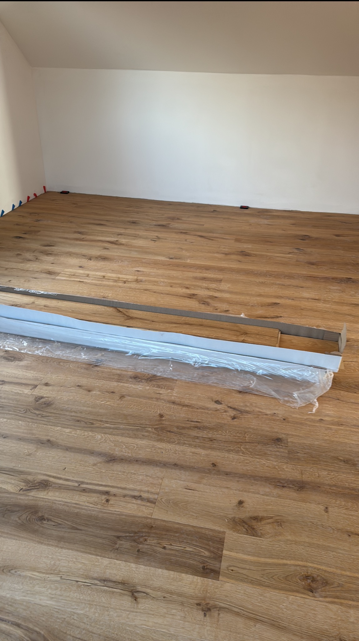 Ułożone panele podłogowe w pokoju na poddaszu, widoczne listwy przypodłogowe w folii i kliny dystansowe przy ścianach. Jasne panele imitujące drewno.