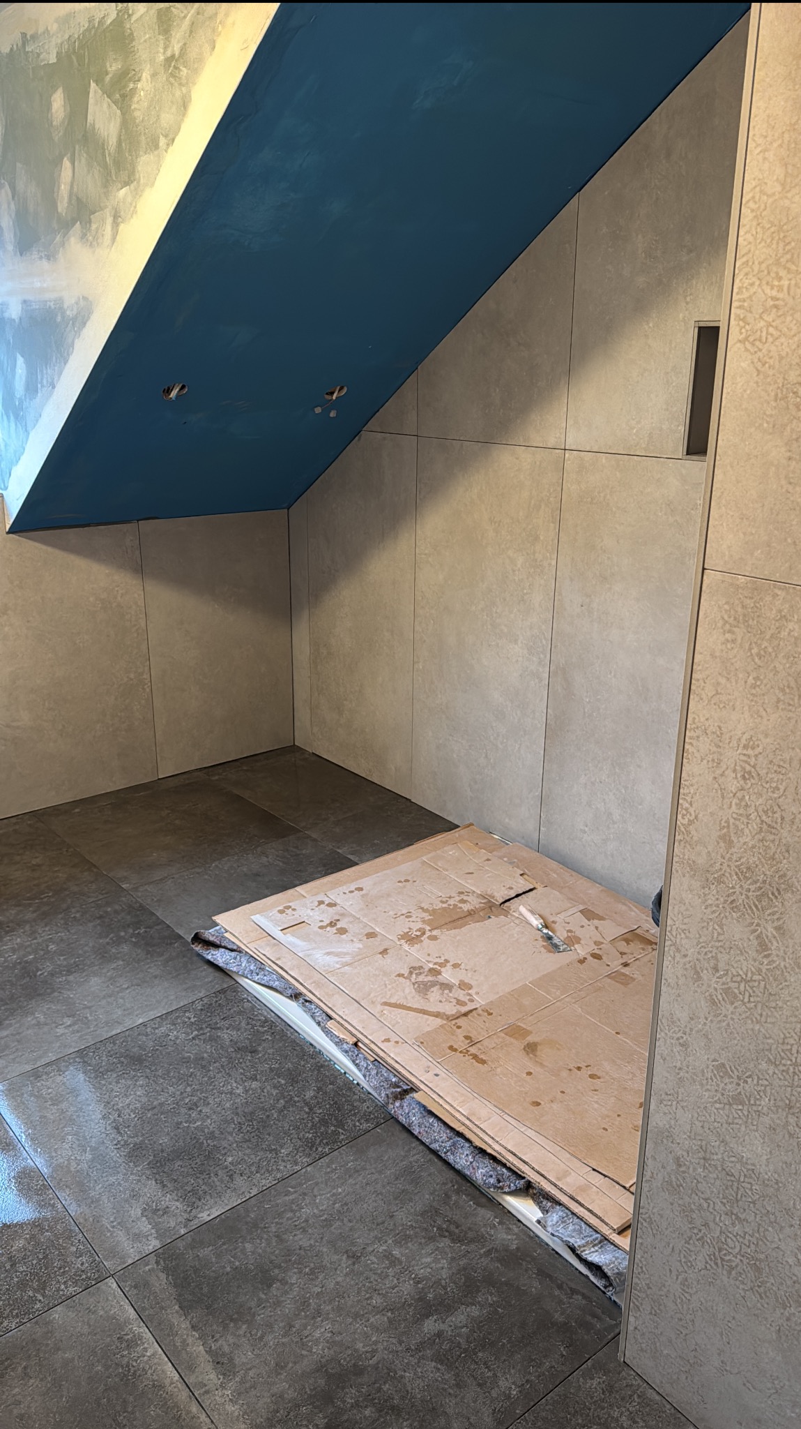 Wykończona łazienka w Jelnej z szarymi płytkami na ścianach i podłodze. Sufit pomalowany na niebiesko. Na podłodze kartony zabezpieczające.