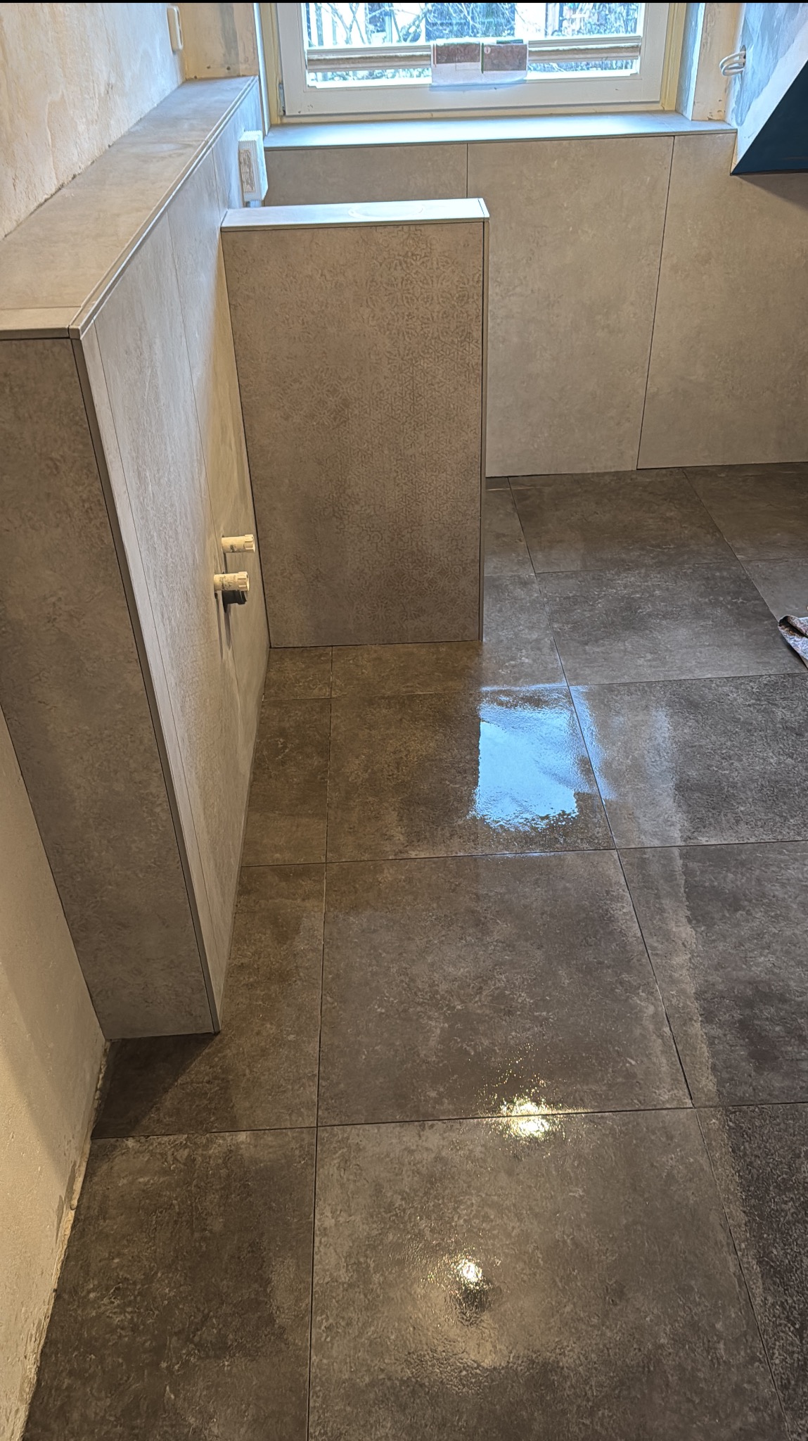 Wykończona łazienka z szarymi płytkami na podłodze i ścianach, widoczne przyłącza wodne. Minimalistyczny design, świeżo po umyciu podłogi, odbicie światła w płytkach.