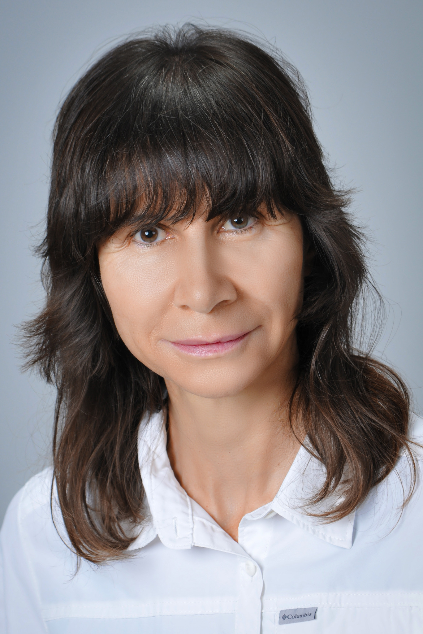 Portret kobiety o brązowych włosach i łagodnym uśmiechu, ubrana w białą koszulę na szarym tle. Profesjonalny wizerunek terapeuty medycyny naturalnej.