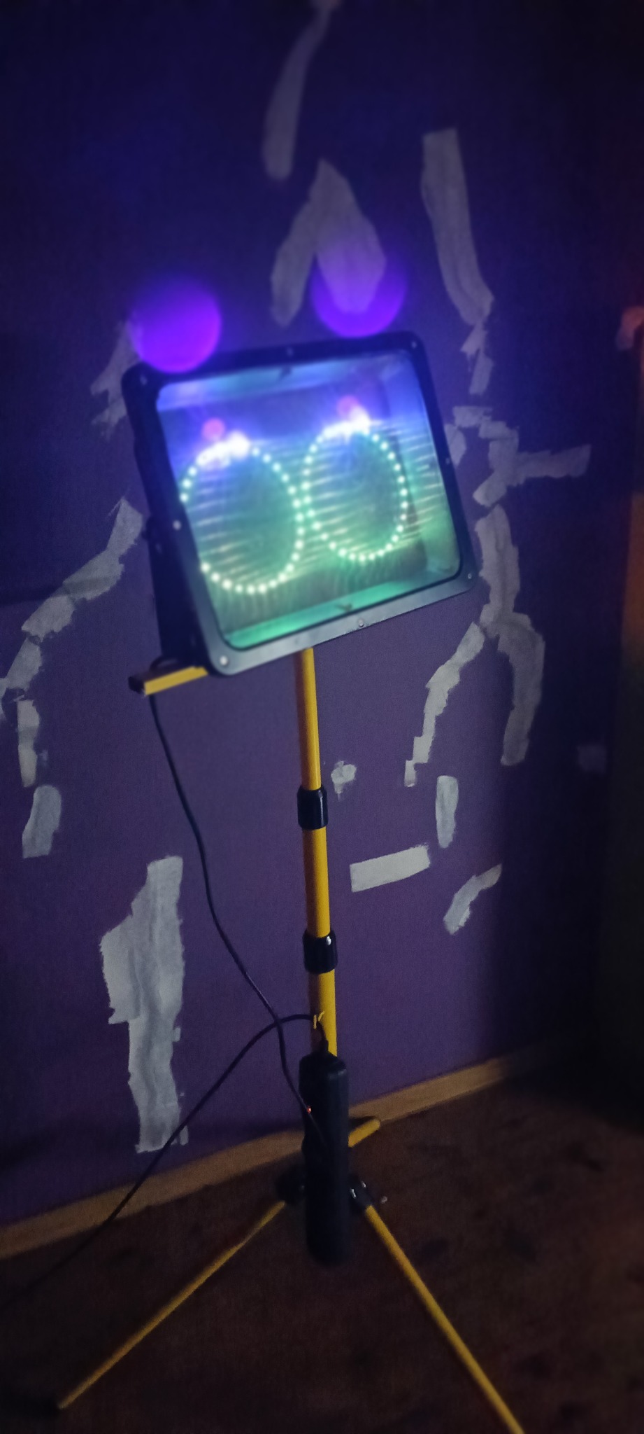 Lampa UV na statywie, emitująca światło w kształcie okręgów, używana do wykrywania zanieczyszczeń lub śladów biologicznych na fioletowej ścianie.