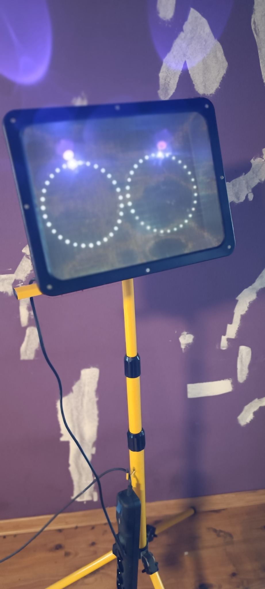 Lampa owadobójcza na żółtym statywie, emitująca światło w kształcie okręgów, ustawiona w pomieszczeniu z fioletową ścianą i śladami po tapecie.