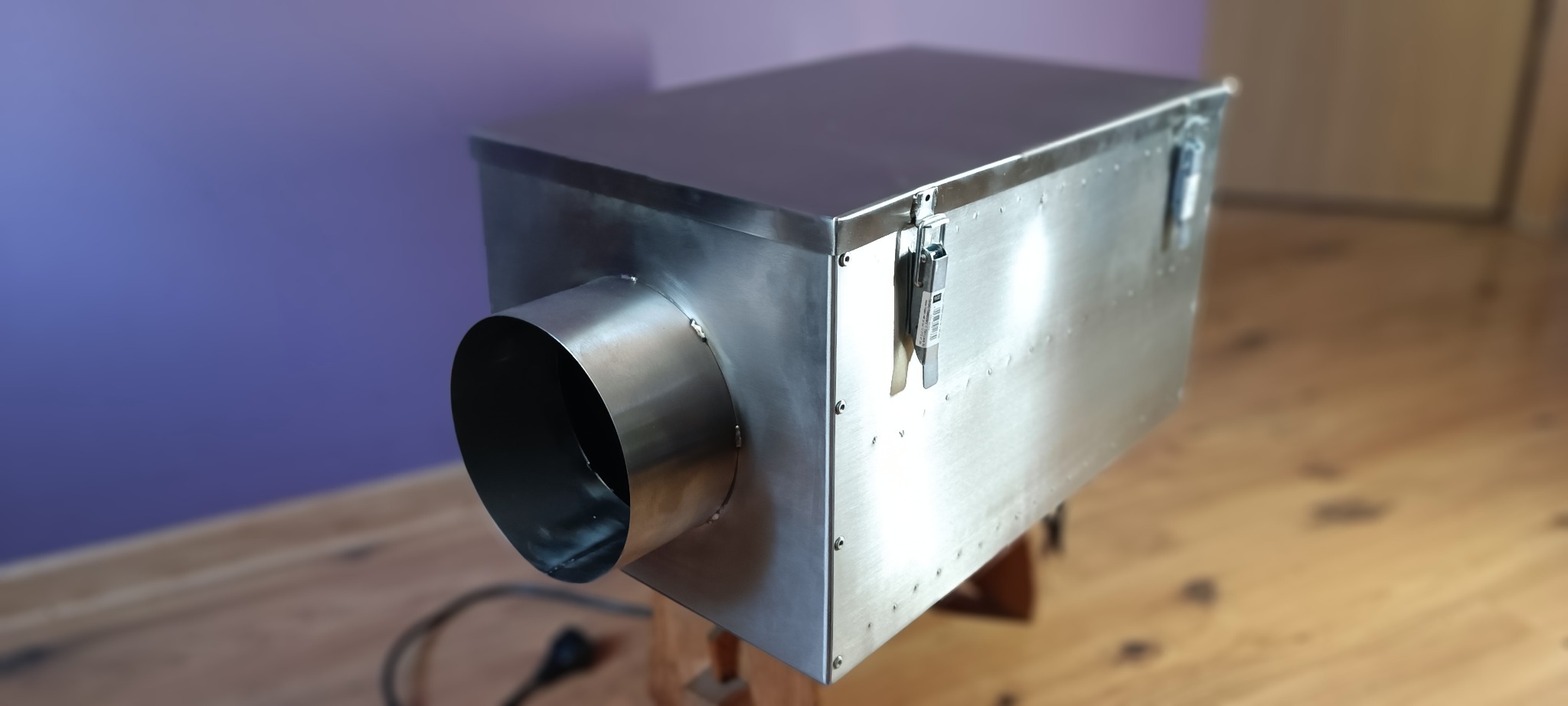 Metalowa skrzynka z okrągłym wlotem, prawdopodobnie urządzenie do zamgławiania, stoi na drewnianym podwyższeniu. Widoczny kabel zasilający i fioletowa ściana w tle.