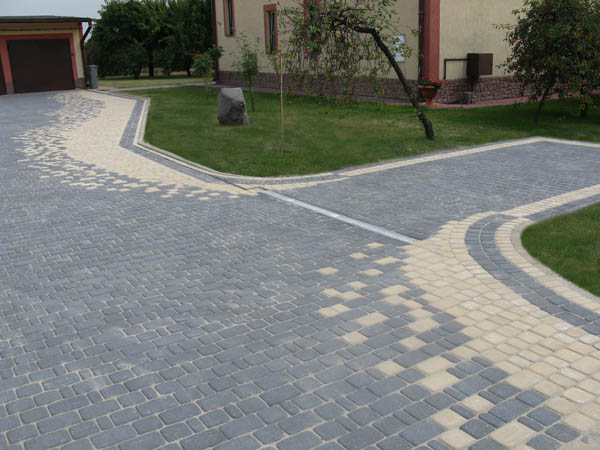 Ułożona kostka brukowa w dwóch odcieniach szarości i beżu tworząca wzór na podjeździe i chodniku przed domem z zielonym trawnikiem i drzewem.