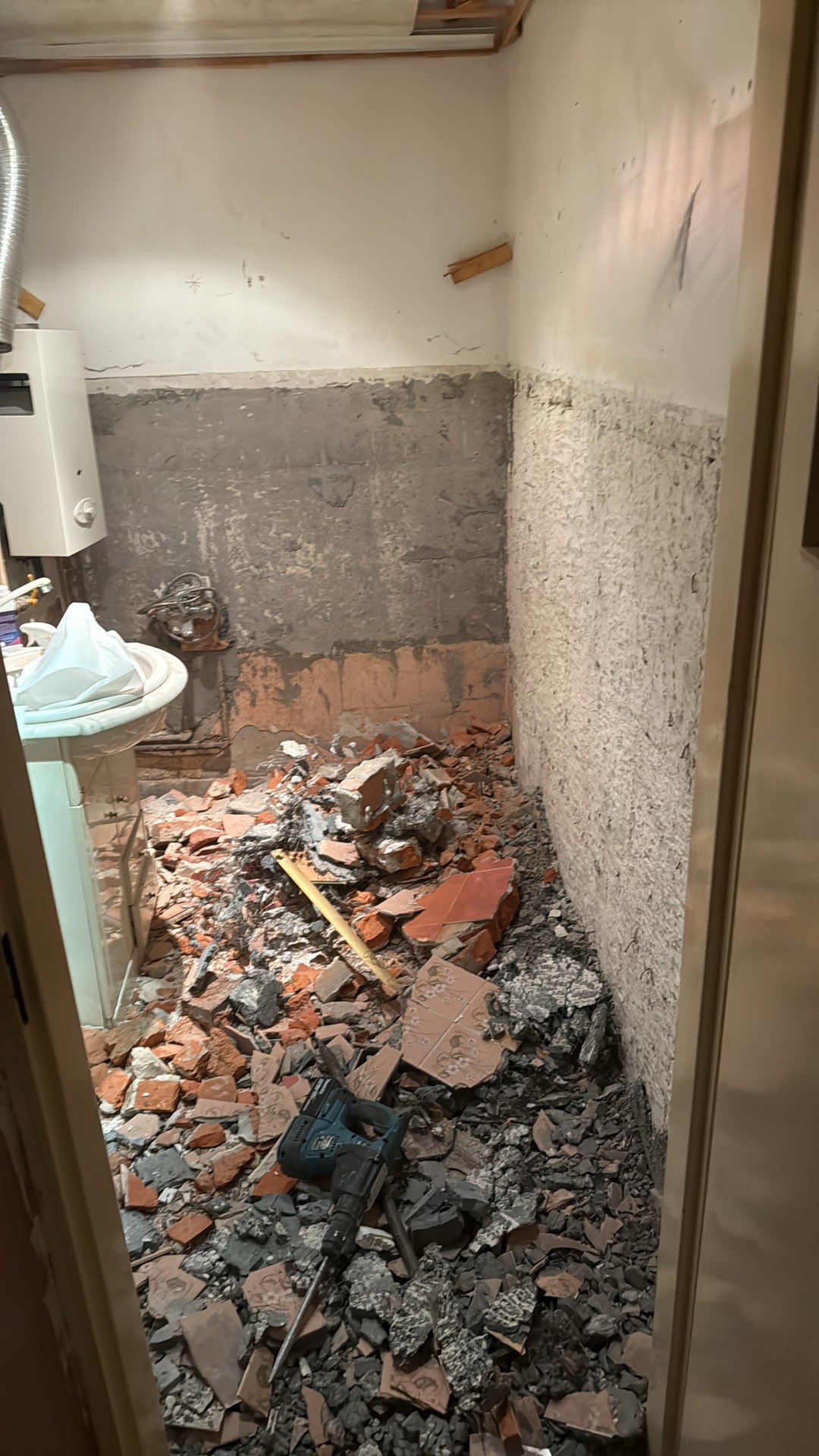 Łazienka w trakcie demontażu płytek, widoczne gruz, odłupane fragmenty ścian i narzędzie do wyburzania. Częściowo zdemontowana umywalka.
