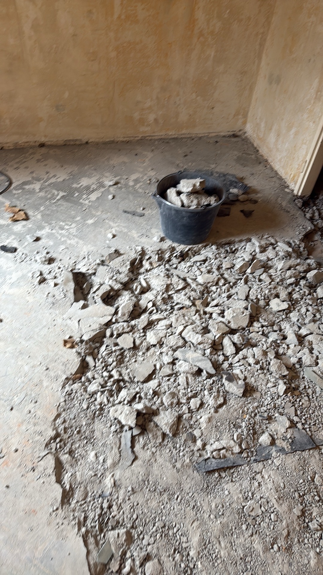 Fragment podłogi po skuciu w pomieszczeniu. Gruz i wiadro z resztkami. Widoczne ślady po usuniętej warstwie podłogi. Ściany w tle. Prace remontowe w toku.