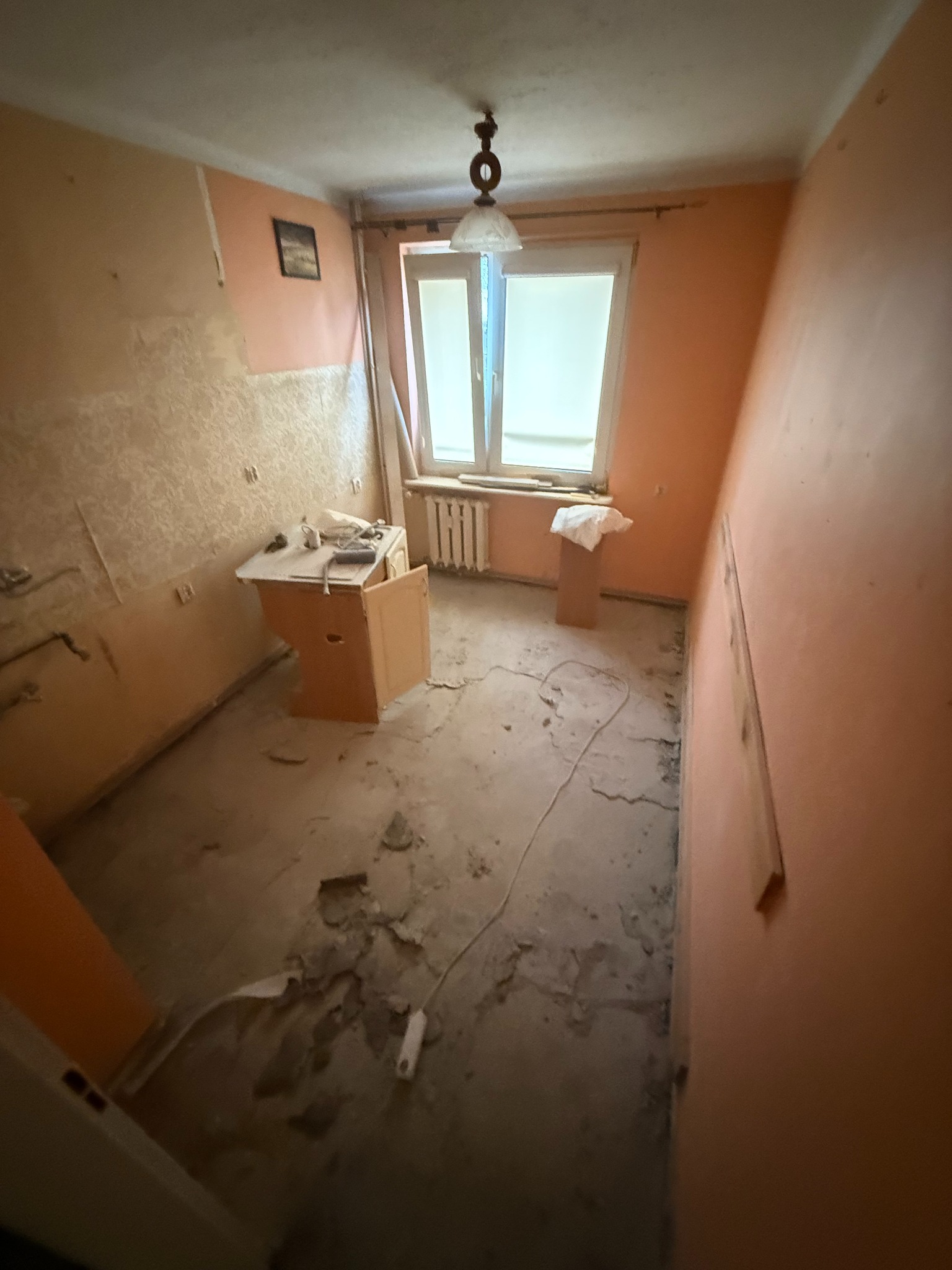 Pokój przed remontem: zerwana tapeta, odsłonięta podłoga, stare okno. Widoczny stolik i lampa sufitowa. Pomieszczenie w trakcie przygotowania do prac remontowych.