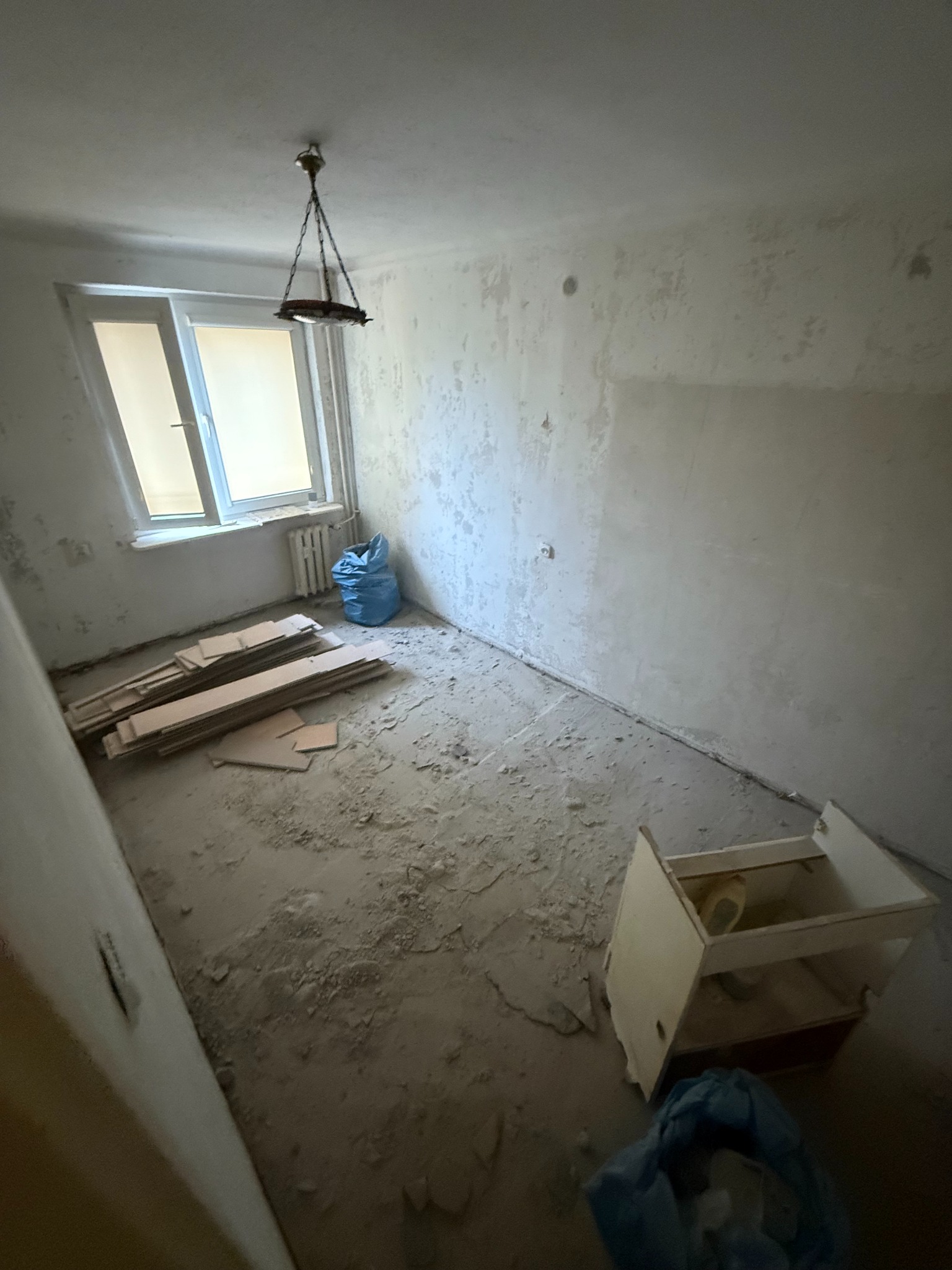 Pokój w trakcie remontu: zdarte tapety, resztki podłogi, worki na śmieci, odsłonięty grzejnik pod oknem, wisząca lampa. Przygotowanie do dalszych prac wykończeniowych.