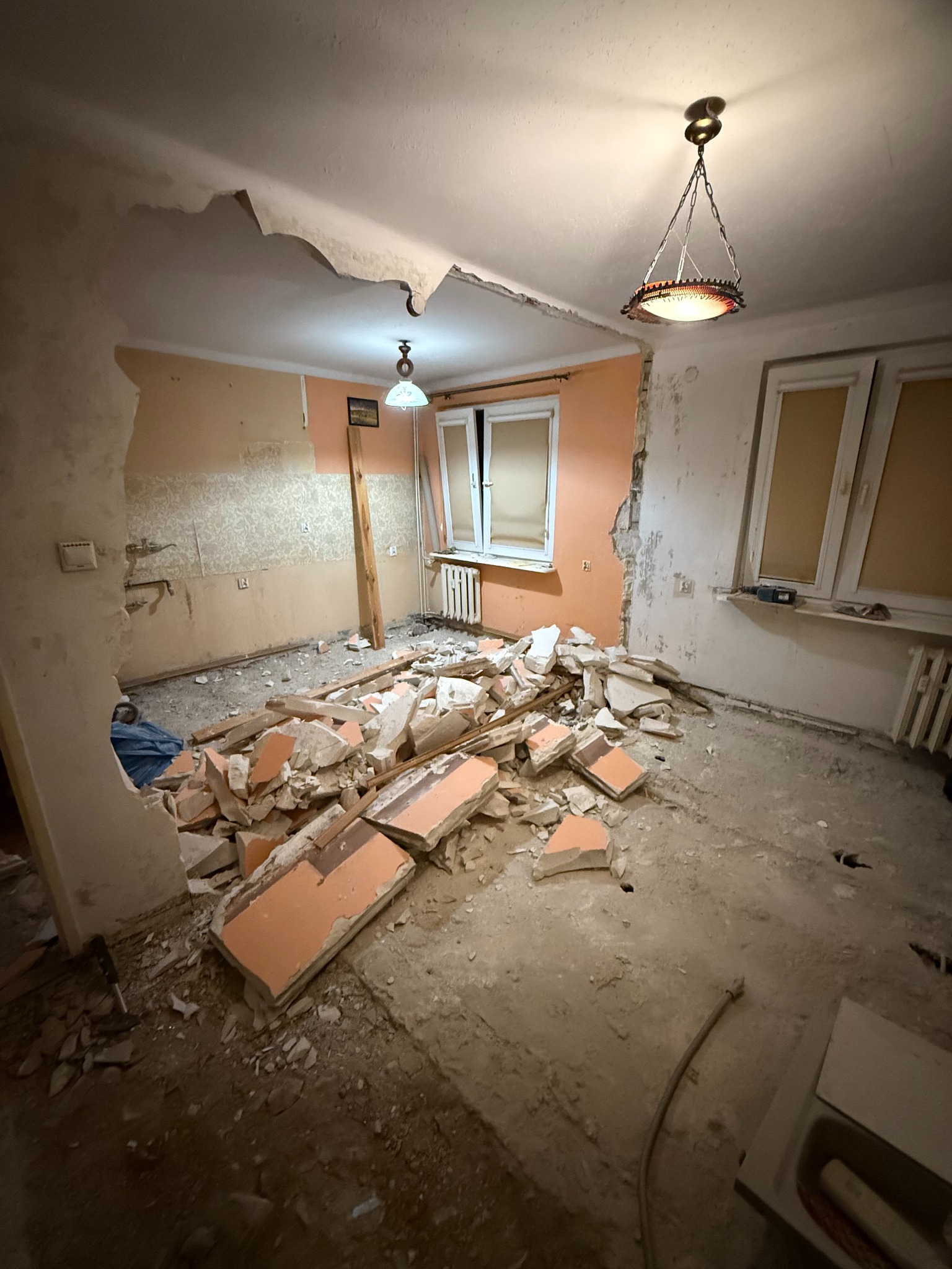 Pokój w trakcie wyburzania ścian, widoczne gruzy, odsłonięte fragmenty ścian i instalacji. Charakterystyczne lampy wiszące. Prace wyburzeniowe w mieszkaniu.