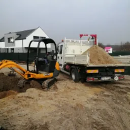 Mała koparka JCB 8018 obok białej ciężarówki z wywrotką pełną piasku na placu budowy; w tle dom z szarą elewacją i różowy domek narzędziowy.