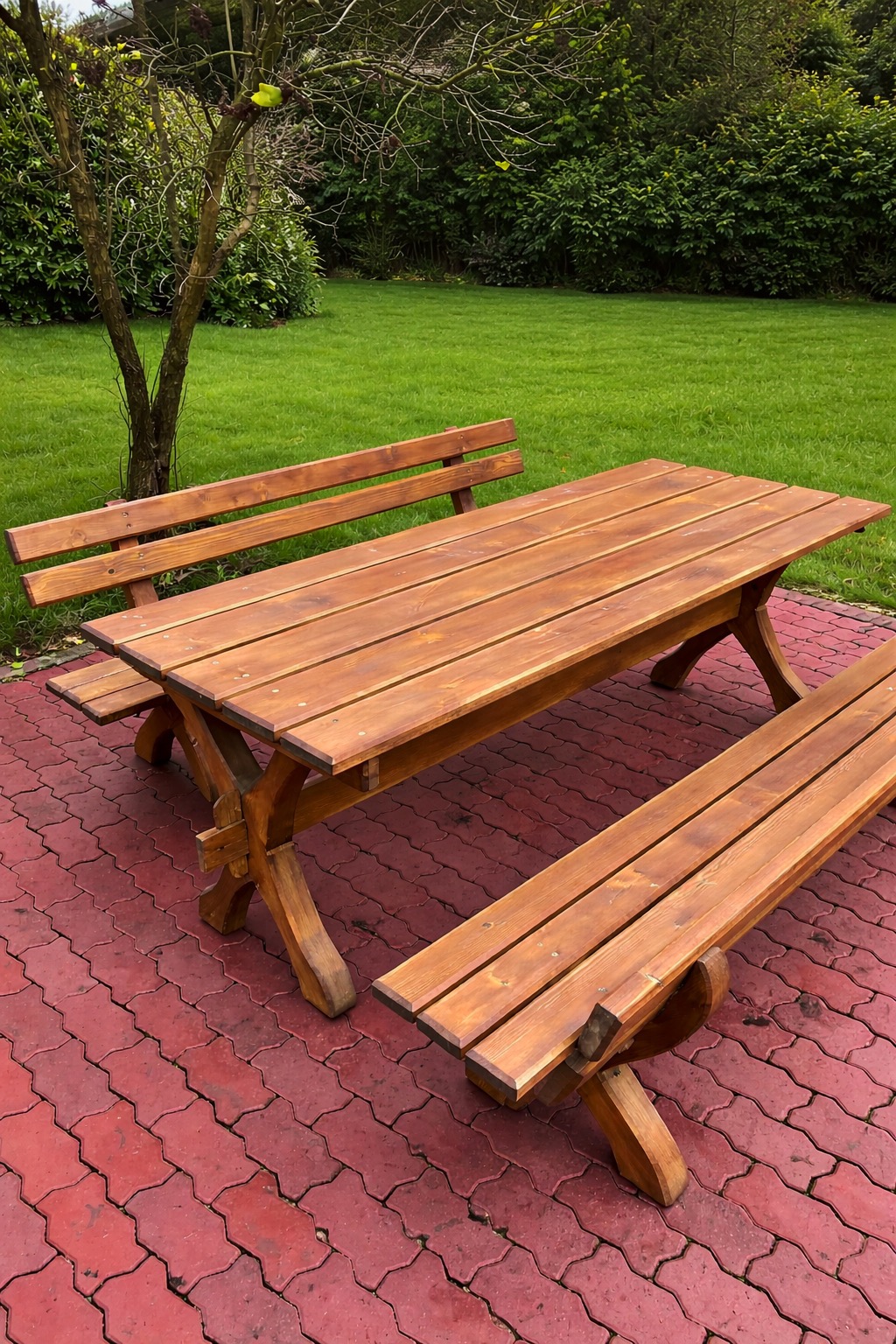 Drewniany stół piknikowy z ławkami na czerwonej kostce brukowej, otoczony zielenią trawnika i drzew. Widoczne detale konstrukcji stołu i ławek.