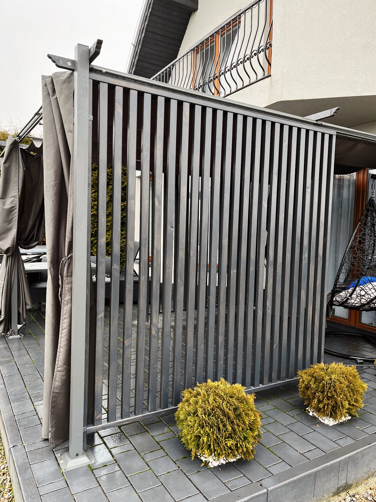 Nowoczesna pergola z szarymi lamelami i zasłonami, uzupełniona o kuliste tuje na tle domu z balkonem. Minimalistyczny design i funkcjonalność w jednym.