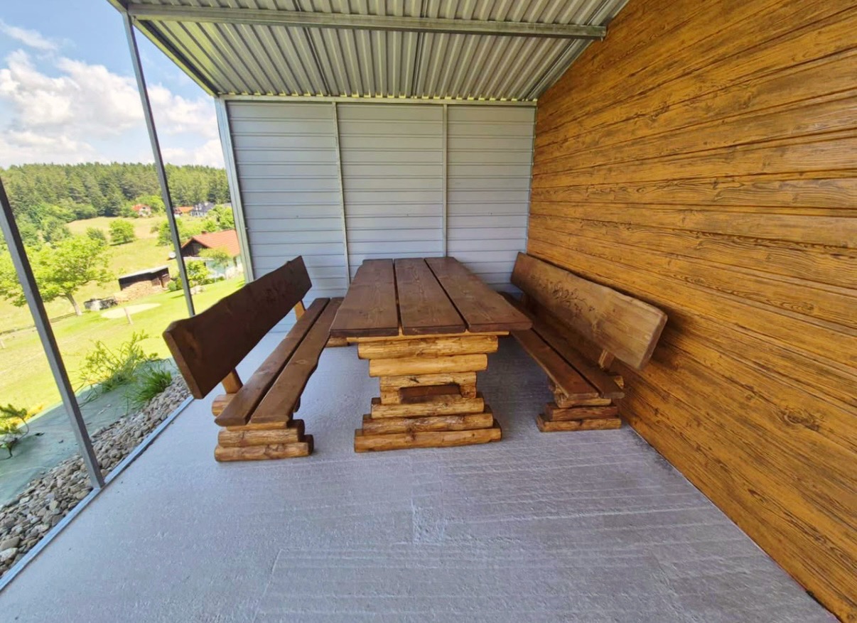 Rustykalny zestaw mebli ogrodowych z bali: stół i ławki pod zadaszeniem, z widokiem na zielony krajobraz. Drewno w ciepłych odcieniach brązu.