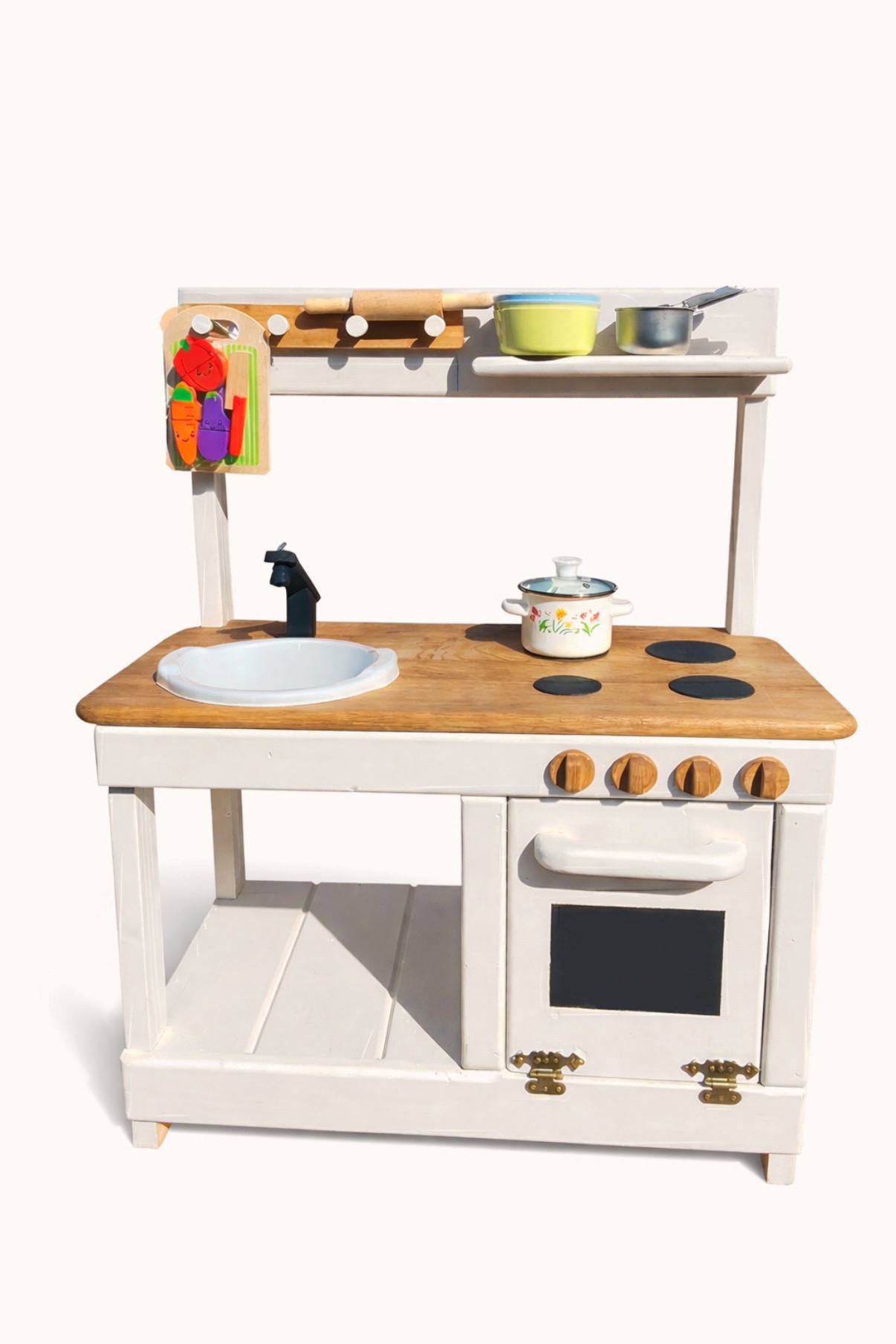 Drewniana kuchnia zabawkowa w stylu rustykalnym, z białymi elementami i blatem z jasnego drewna. Widoczne akcesoria: garnki, zlew, imitacja palników i piekarnika.