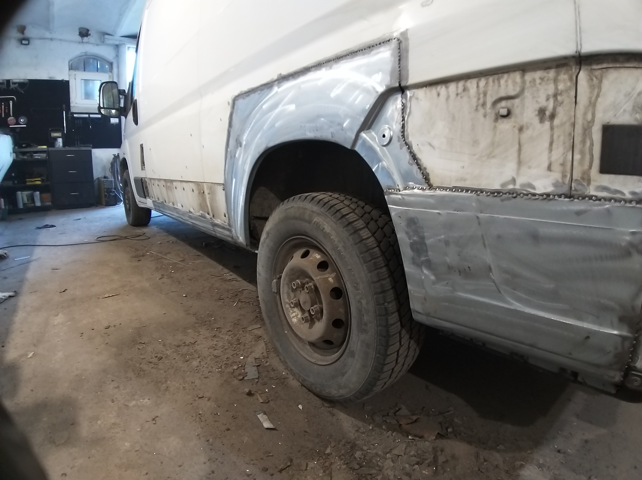 Biały samochód dostawczy w trakcie naprawy blacharskiej. Widoczne nowe poszycie nadkola i progu, po spawaniu. Warsztat z narzędziami w tle.