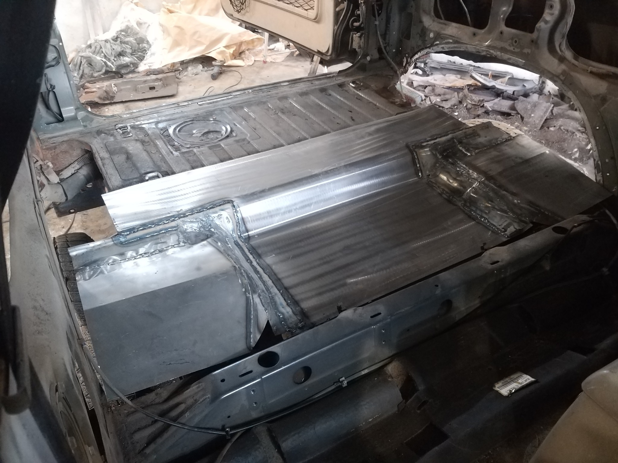 Wnętrze samochodu w trakcie naprawy podłogi. Widoczne nowe, metalowe elementy podłogi, częściowo już wspawane, surowe spawy. Prace blacharskie w toku.