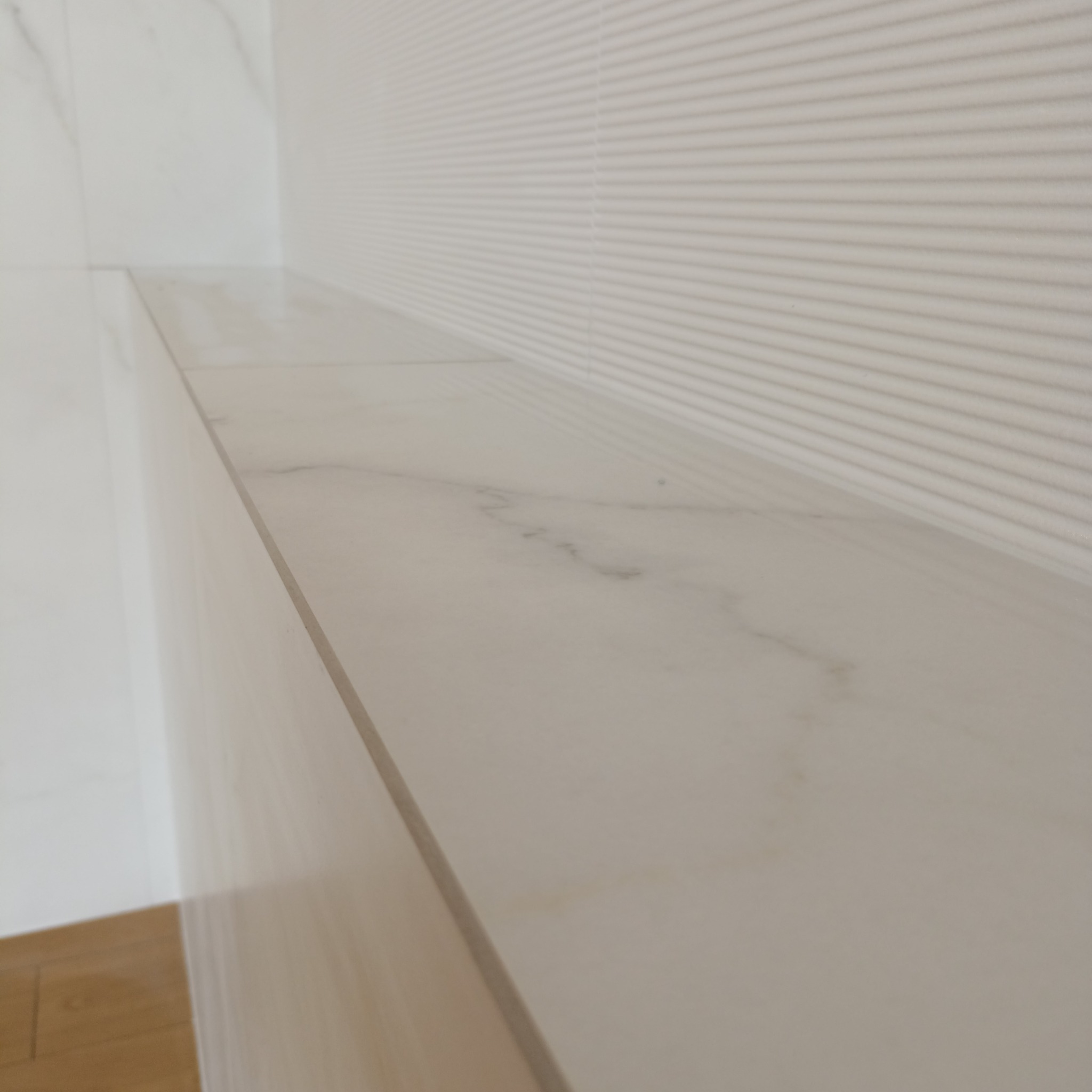 Półka z płytek imitujących marmur, zamontowana w rogu łazienki z dekoracyjną, prążkowaną ścianą. Widoczny fragment podłogi z drewnopodobnych płytek.