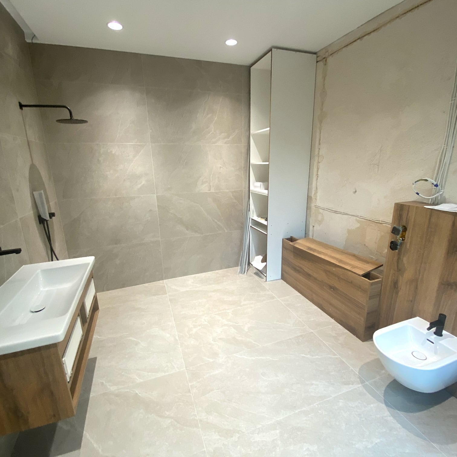 Jasna łazienka w trakcie wykańczania: biała umywalka i bidet, drewniane szafki, beżowe płytki, surowa ściana. Widoczne elementy instalacji.