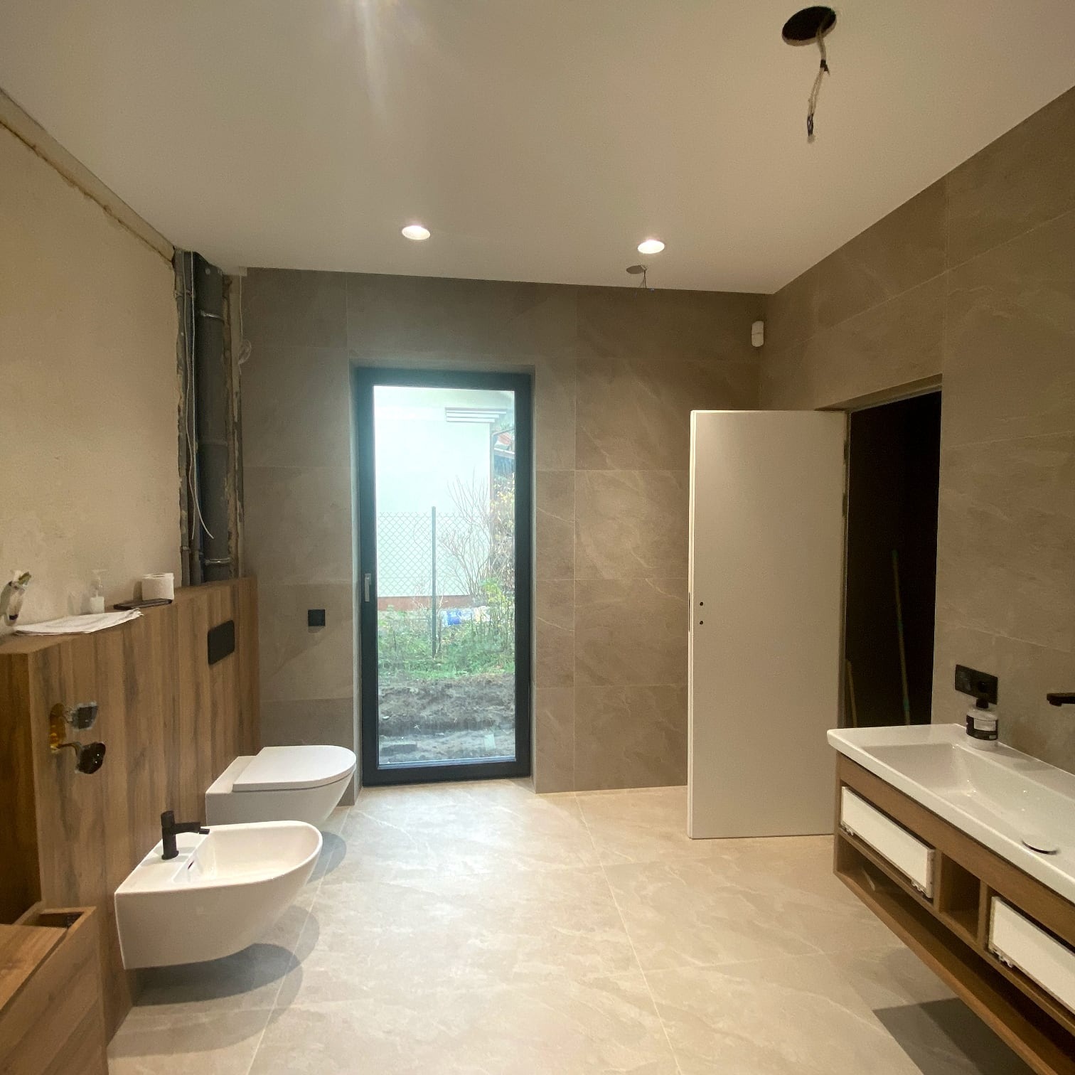 Jasna łazienka z beżowymi płytkami, drewnianymi akcentami i nowoczesną armaturą. Widoczne WC, bidet, umywalka z szafką i okno z widokiem na zewnątrz.