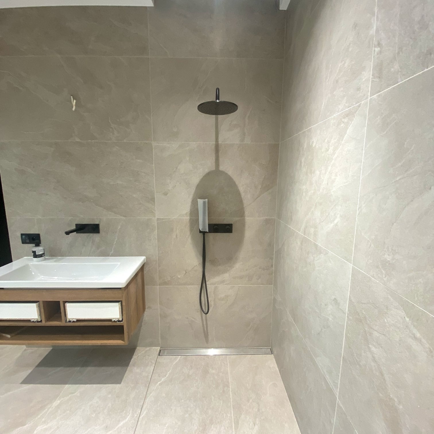 Jasna łazienka z beżowymi płytkami, kabiną prysznicową typu walk-in i wiszącą szafką z umywalką. Minimalistyczny design z czarnymi akcentami.