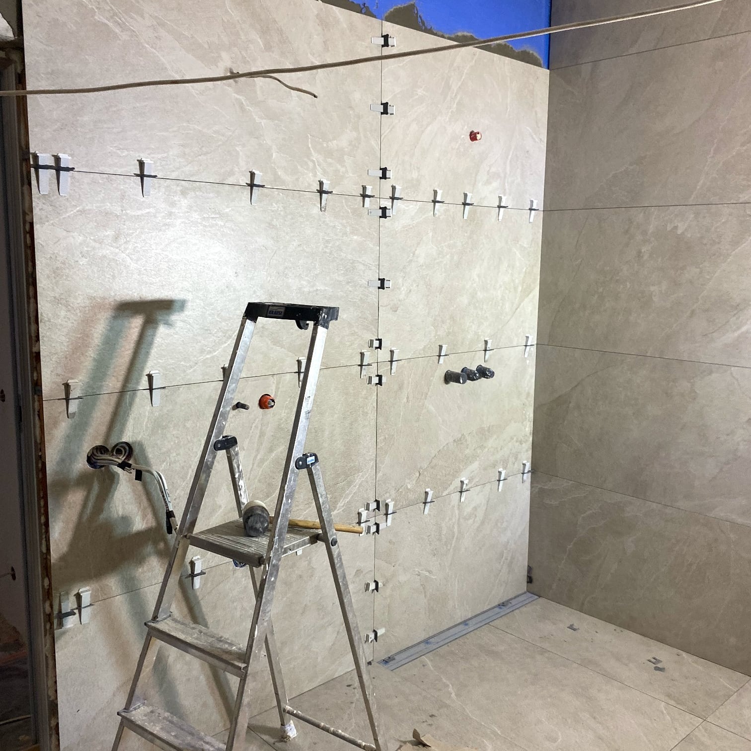 Układanie beżowych płytek w rogu pomieszczenia, widoczne krzyżyki dystansowe, poziomica i drabina. Prace glazurnicze w trakcie realizacji.