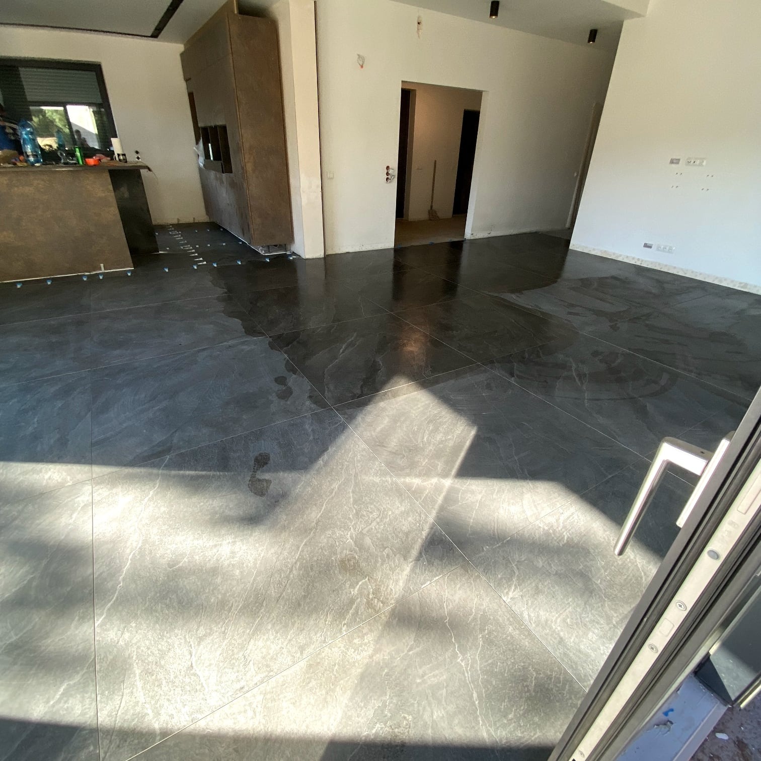 Połyskująca, ciemna podłoga z płytek imitujących marmur w przestronnym wnętrzu. Widoczne krzyżyki dystansowe przy zabudowie kuchennej. Wpadające promienie słoneczne.