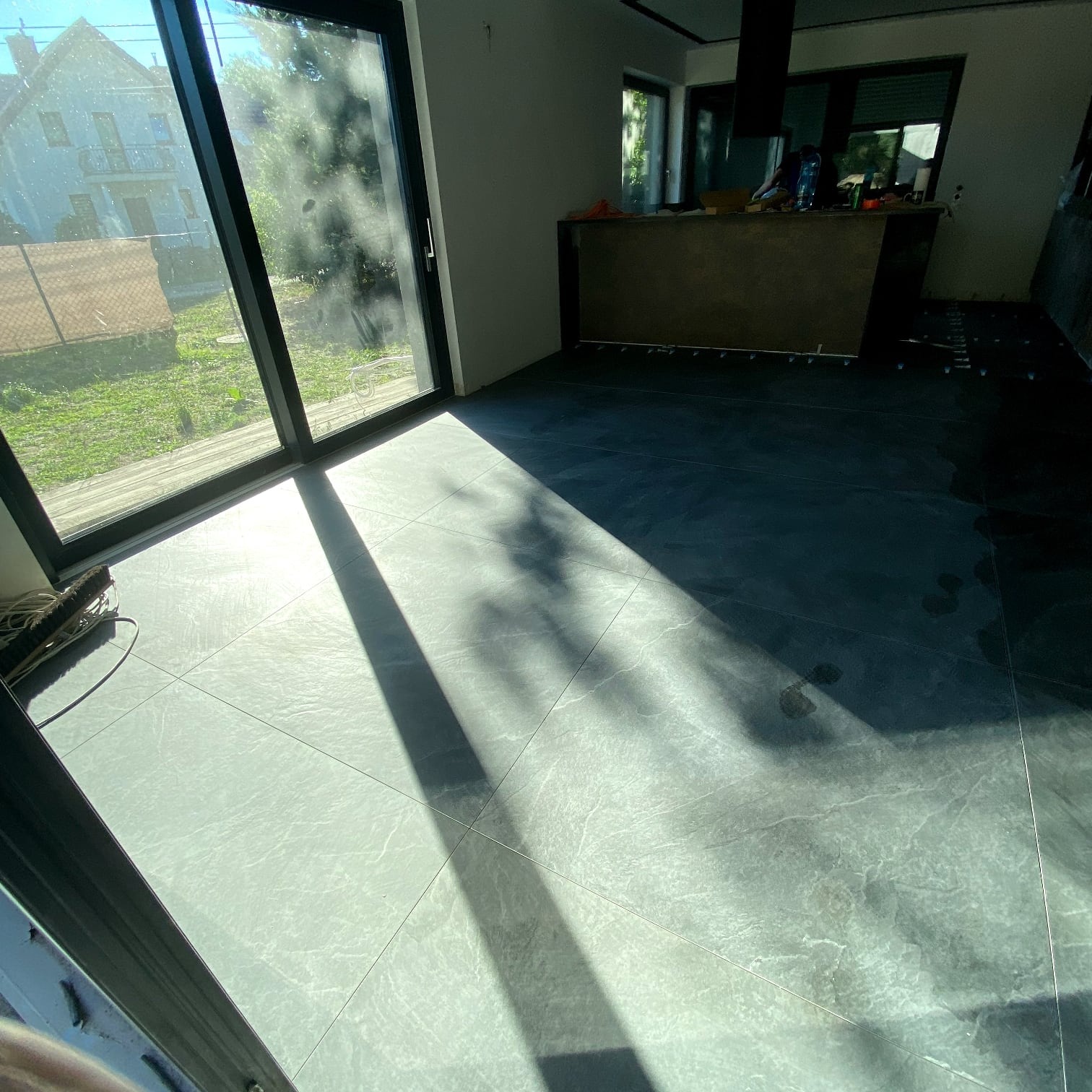 Ułożona, szara podłoga z płytek w jasnym wnętrzu z dużym oknem balkonowym. Widoczne ślady stóp i cień drzewa. Częściowo wykończona kuchnia w tle.