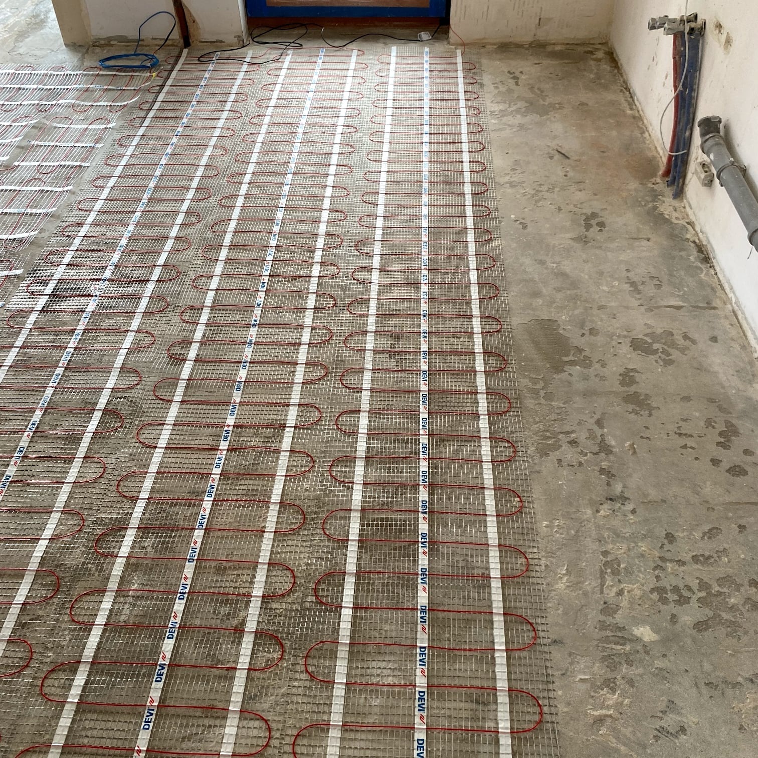 Rozłożona mata grzewcza DEVI na surowym podłożu, przygotowana do wylewki. Widoczne rury instalacyjne przy ścianie. Etap prac przed wykończeniem podłogi.