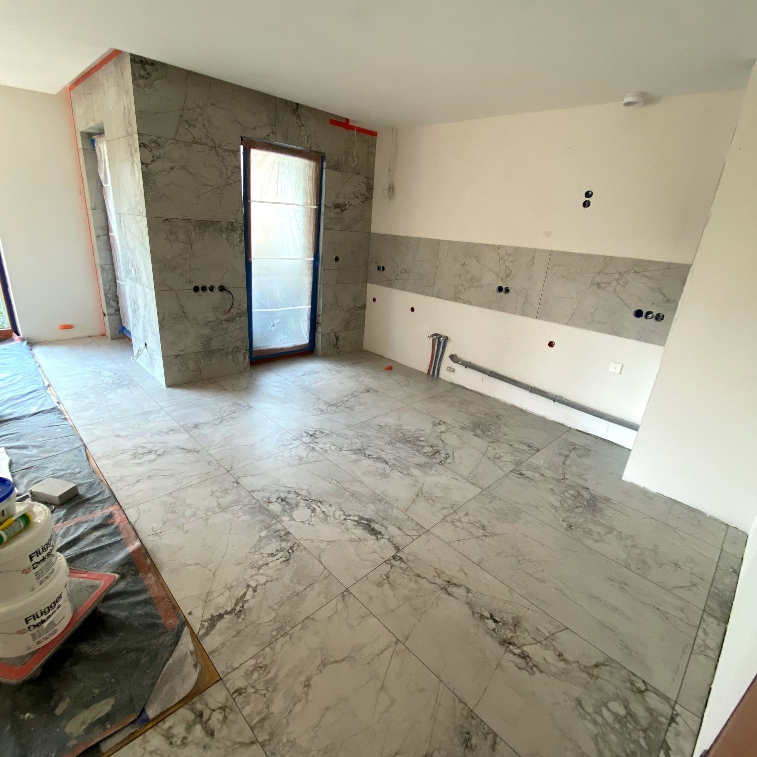 Układanie płytek imitujących marmur na podłodze i ścianie w trakcie prac. Widoczne narzędzia i materiały budowlane. Otwory na gniazdka elektryczne.