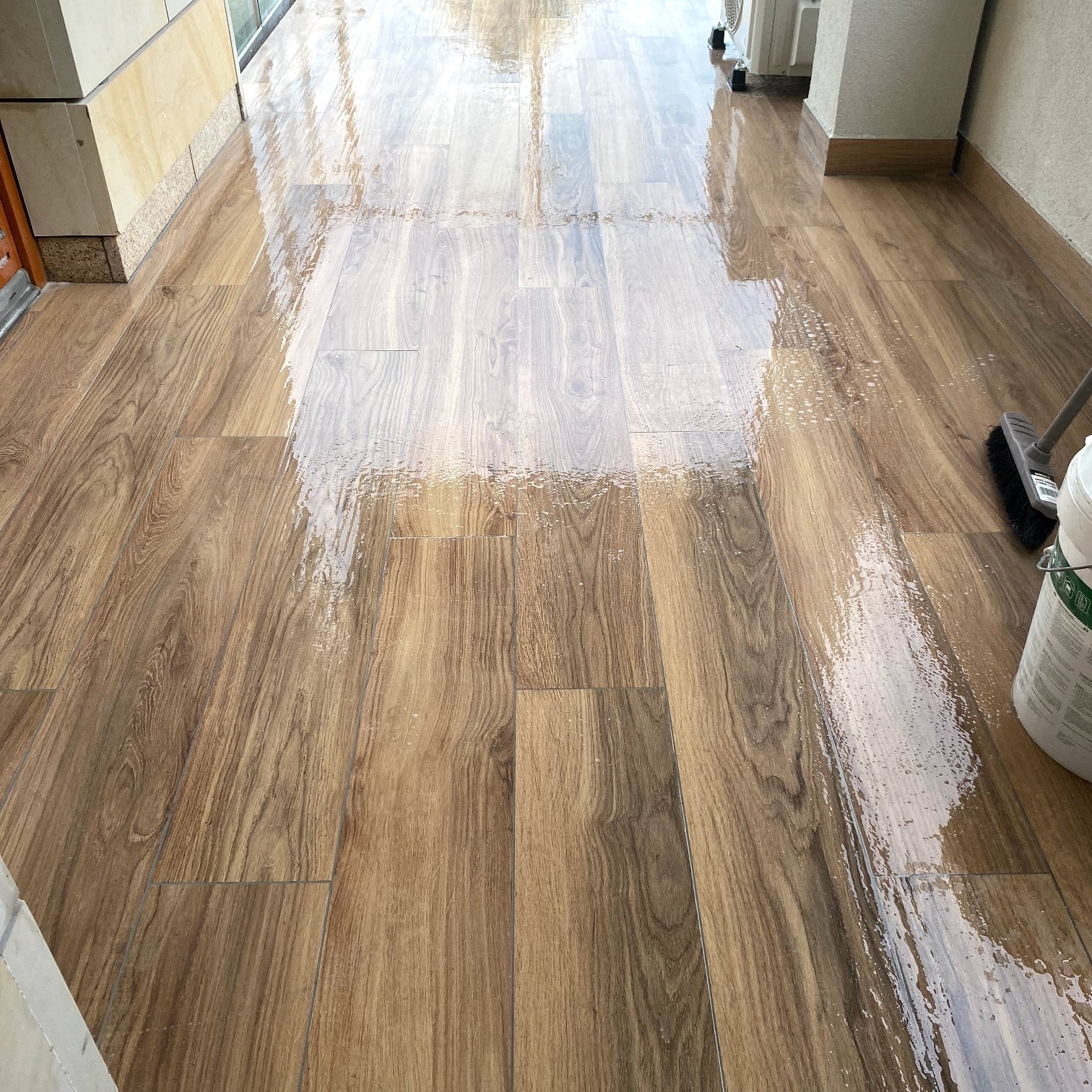 Błyszcząca, drewnopodobna podłoga z płytek po umyciu, z widocznym wiadrem i szczotką w prawym rogu. W tle fragment balkonu i klimatyzator.