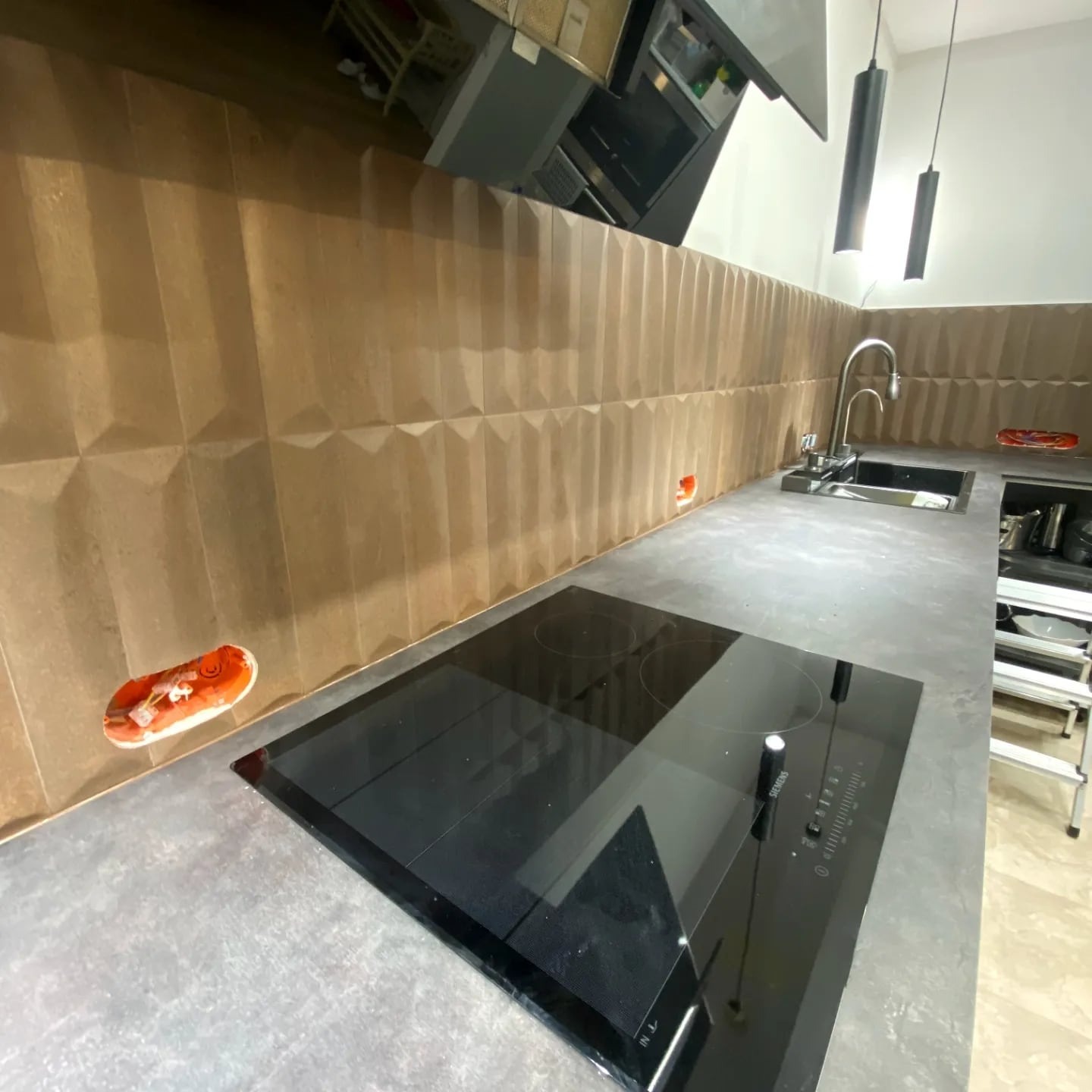 Nowoczesna kuchnia po remoncie: brązowe, trójwymiarowe płytki na ścianie, szary blat, czarna płyta indukcyjna i minimalistyczne lampy wiszące. Widoczne gniazdka elektryczne.