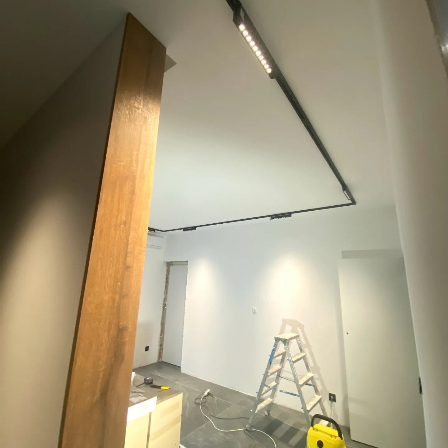 Montaż szynoprzewodów z oświetleniem LED na suficie w jasnym wnętrzu. Widoczna drabina, narzędzia i drewniany element konstrukcyjny. Minimalistyczny design.