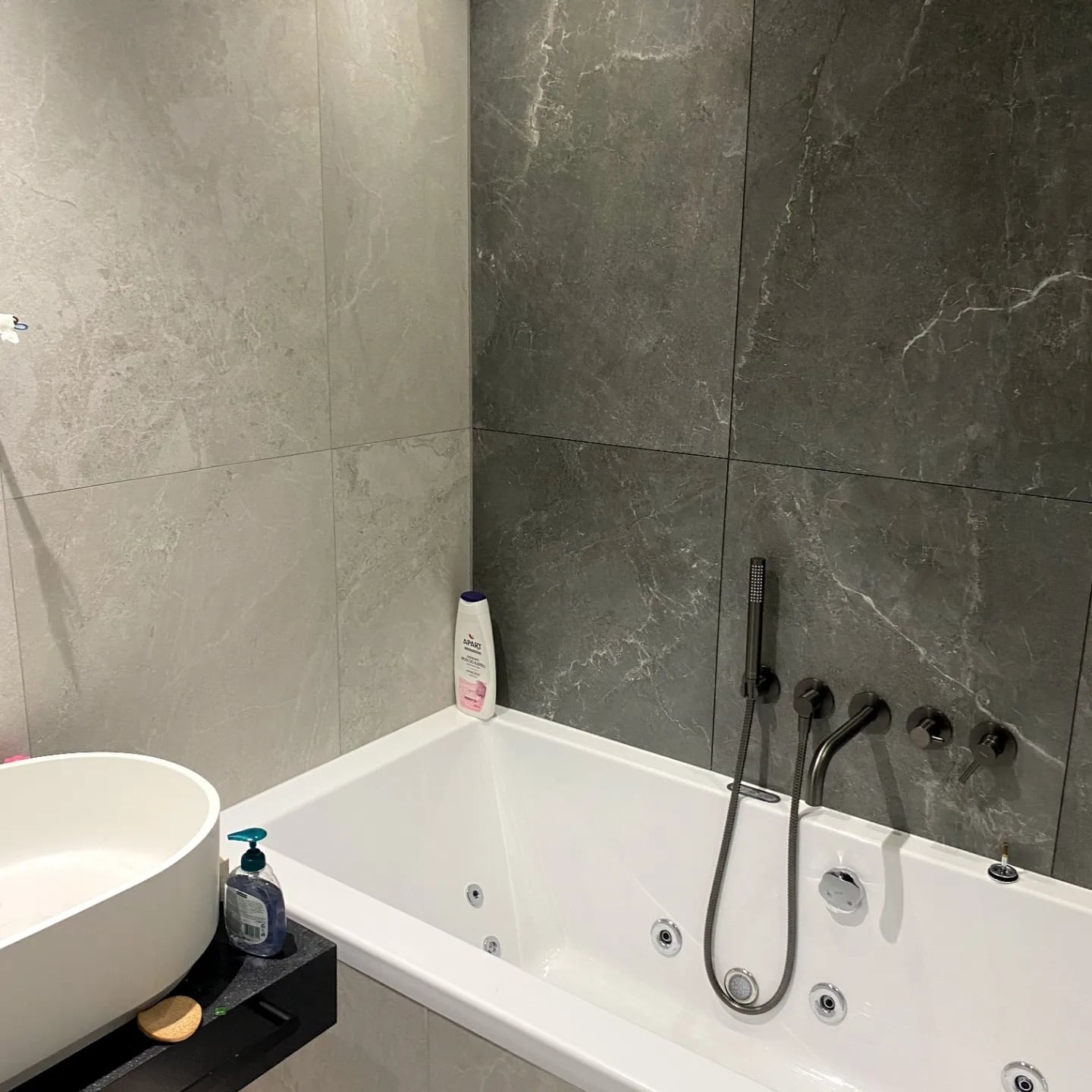 Nowoczesna łazienka z wanną jacuzzi, szare płytki imitujące marmur na ścianach, minimalistyczna armatura w kolorze grafitu i owalna umywalka nablatowa.