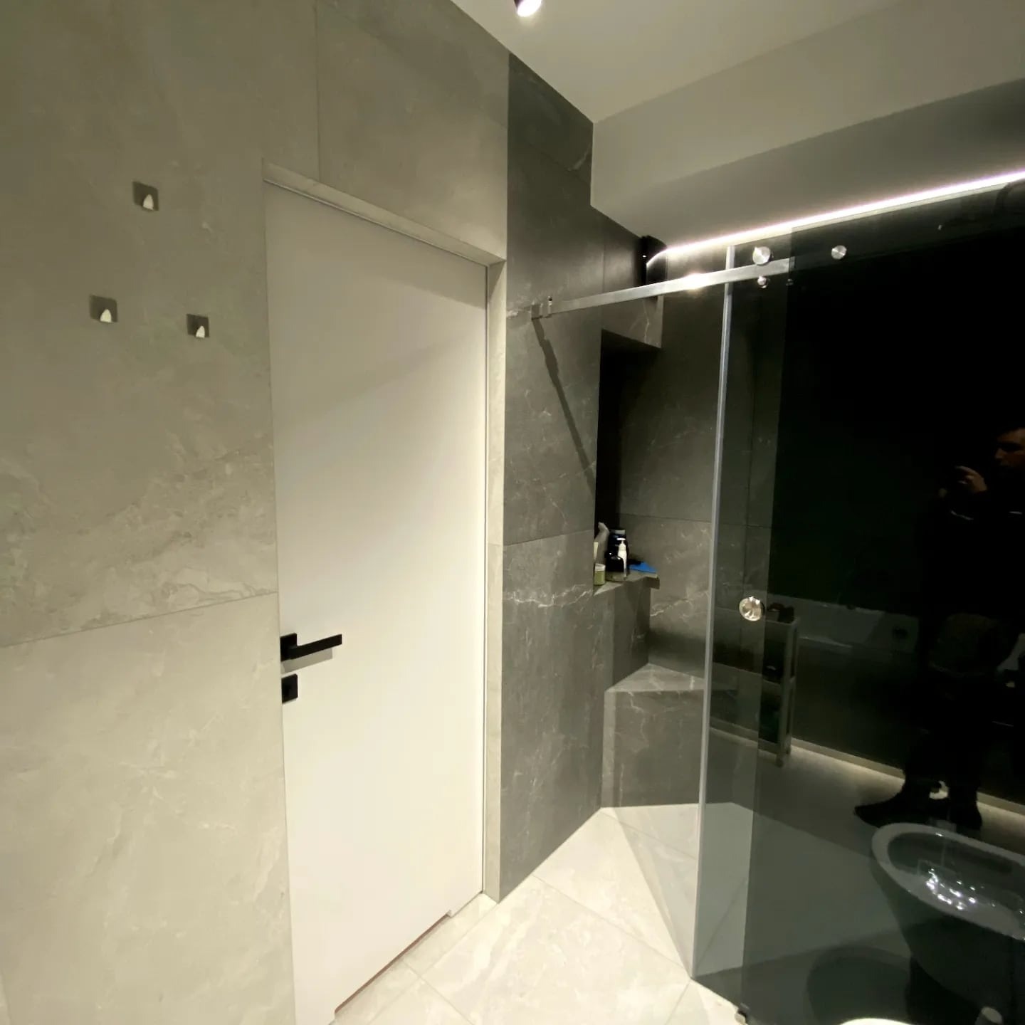 Nowoczesna łazienka z szarymi płytkami imitującymi marmur, białą stolarką drzwiową i szklaną kabiną prysznicową. Minimalistyczne oświetlenie punktowe.