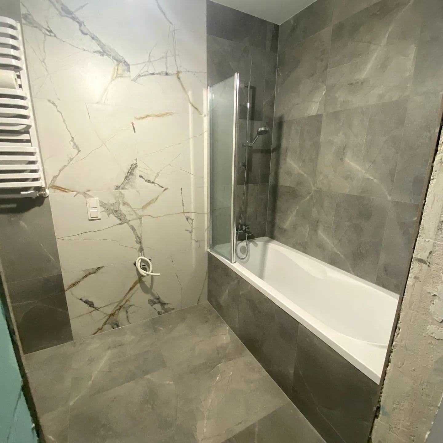 Nowoczesna łazienka z wanną obudowaną szarą płytką imitującą marmur, białą armaturą i grzejnikiem drabinkowym. Ściany wykończone płytkami w dwóch odcieniach szarości.