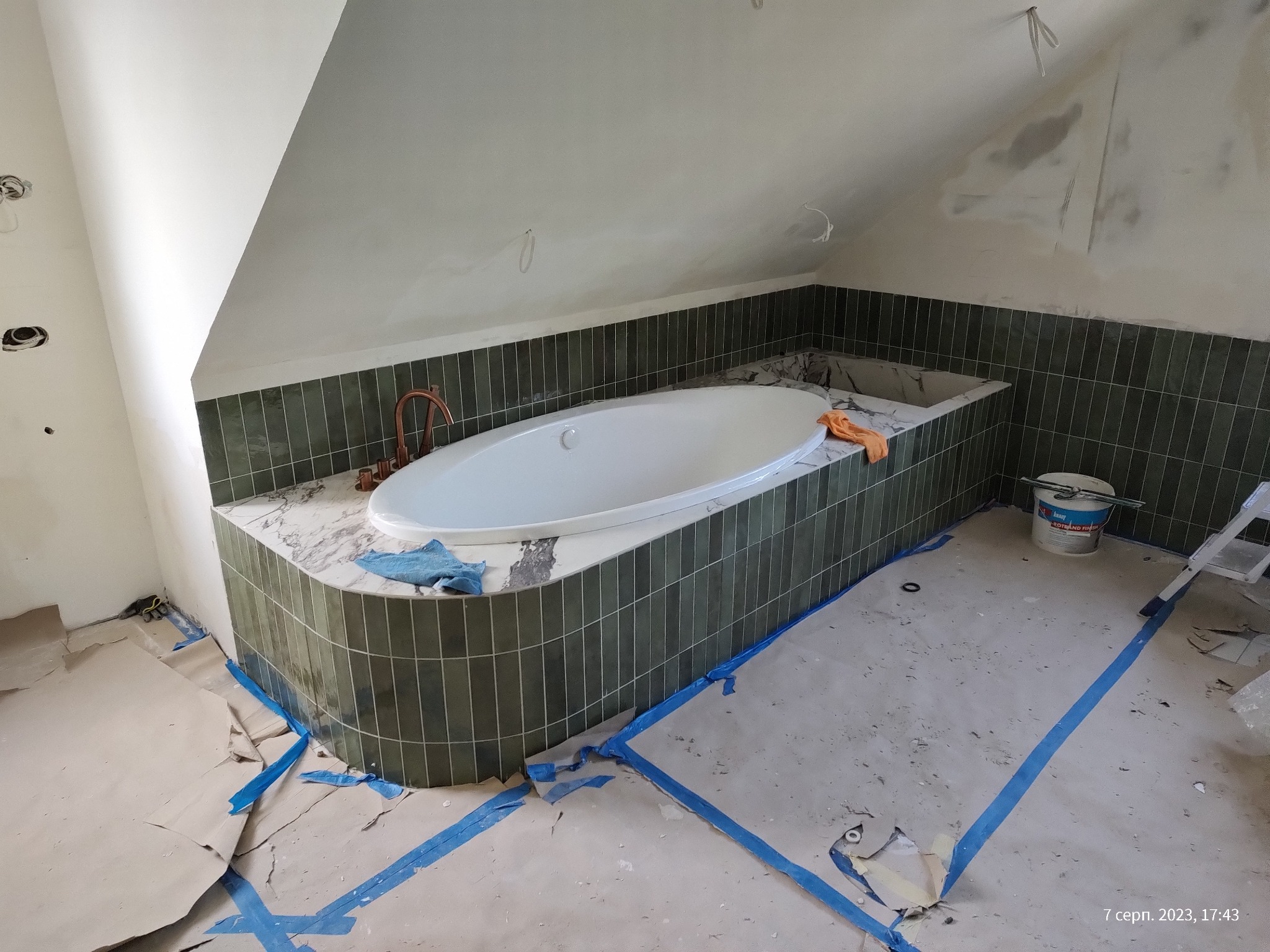 Wnętrze łazienki w trakcie prac wykończeniowych: obudowana wanna z zieloną glazurą, marmurowy blat, zabezpieczona podłoga, widoczne narzędzia i materiały budowlane.