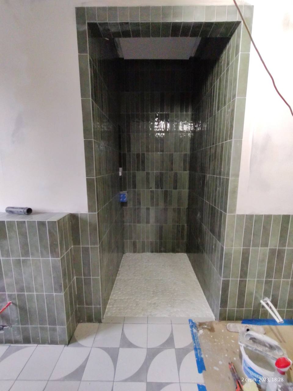 Wnęka prysznicowa z zielonymi, pionowo ułożonymi płytkami. Na podłodze jasne, okrągłe kamyczki. Częściowo widoczne narzędzia i wiadro.