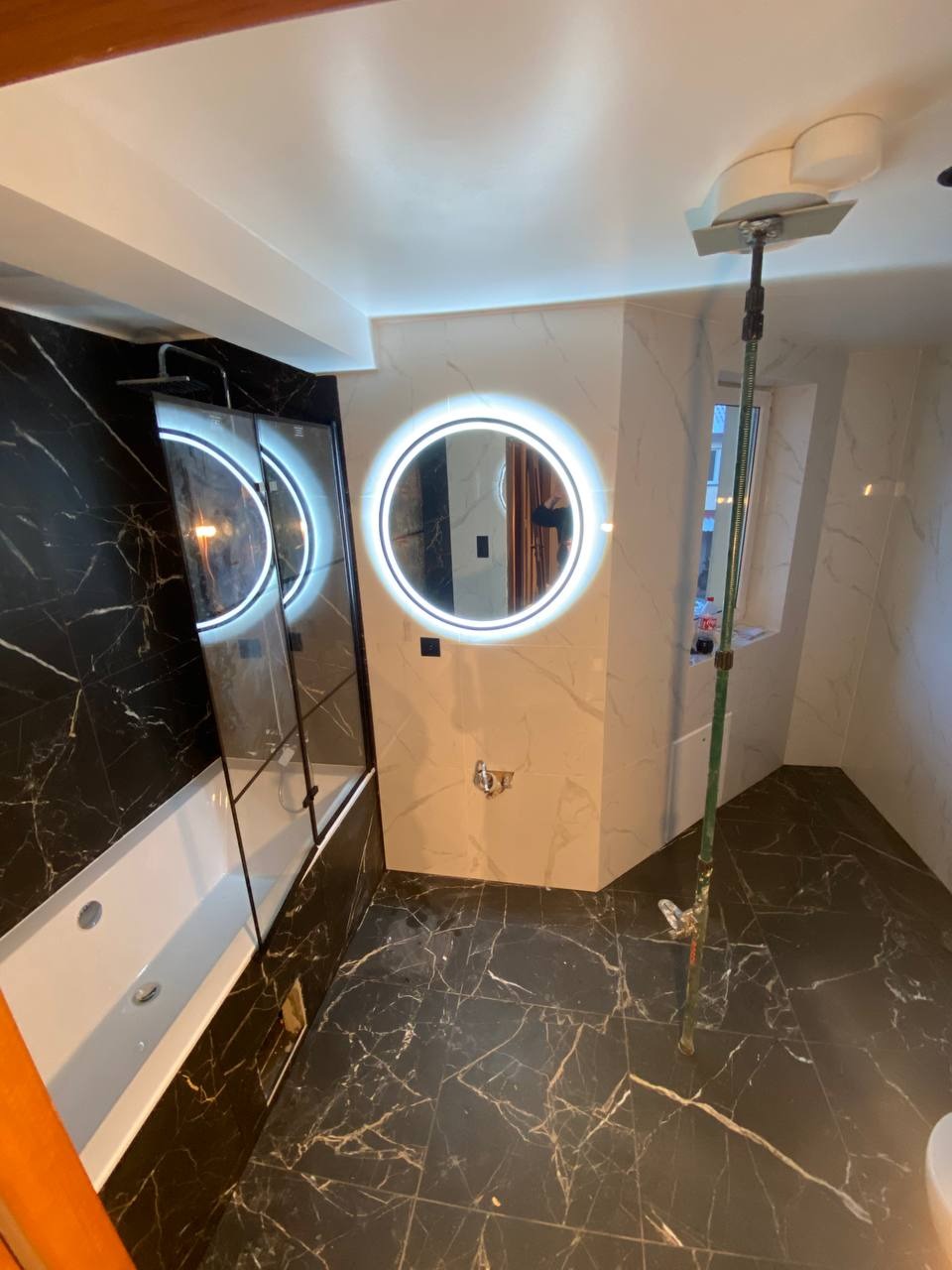 Elegancka łazienka z czarną marmurową podłogą i ścianami, okrągłe lustro z podświetleniem LED, biała wanna z parawanem. Widoczny sprzęt do czyszczenia.