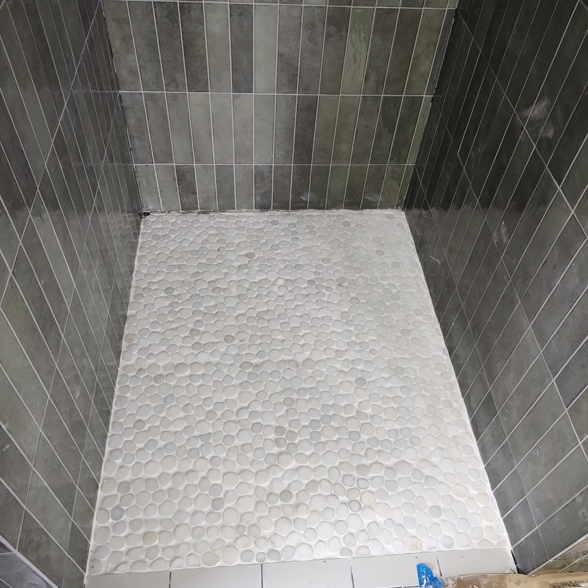 Wykończony brodzik z białych, okrągłych kamyków i szare, pionowe płytki na ścianach. Widok z góry na świeżo położoną mozaikę w kabinie prysznicowej.