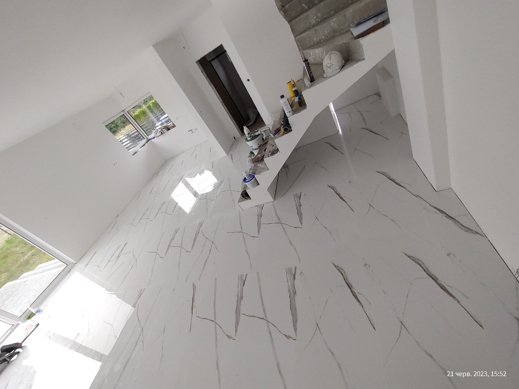 Biała marmurowa podłoga z brązowymi żyłami w jasnym wnętrzu. Widoczne narzędzia i materiały budowlane przy schodach. Duże okno wpuszcza naturalne światło.