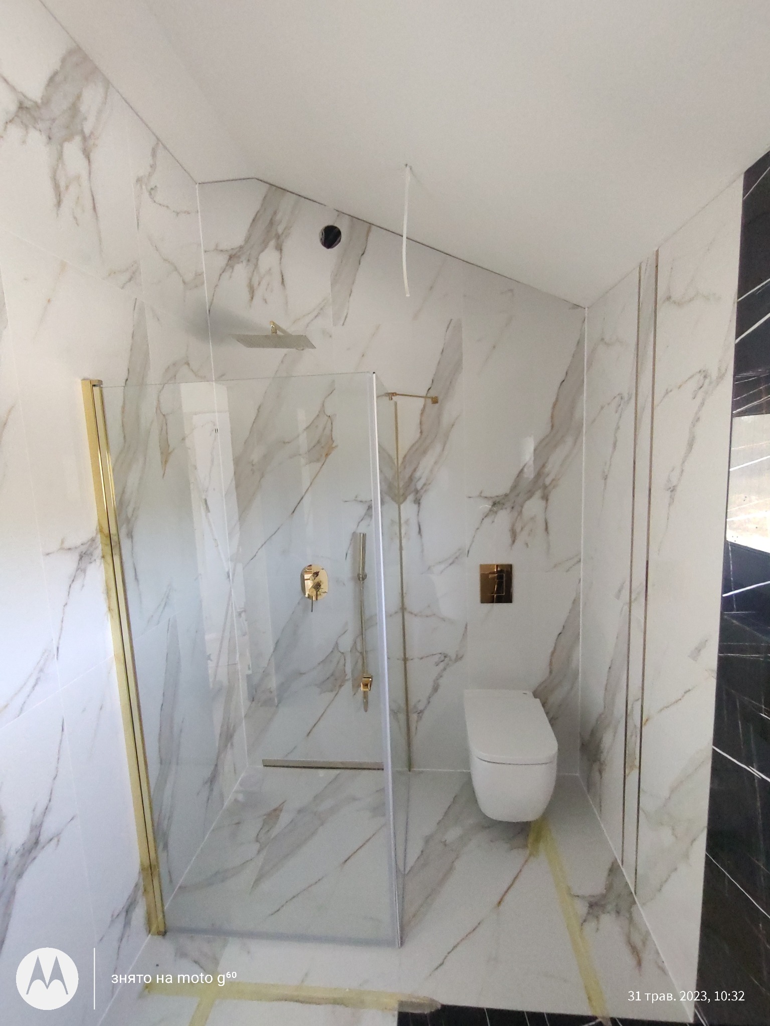 Wykończona łazienka z kabiną prysznicową ze złotymi detalami i marmurowymi płytkami. Widoczna toaleta podwieszana i odpływ liniowy. Nowoczesny design.